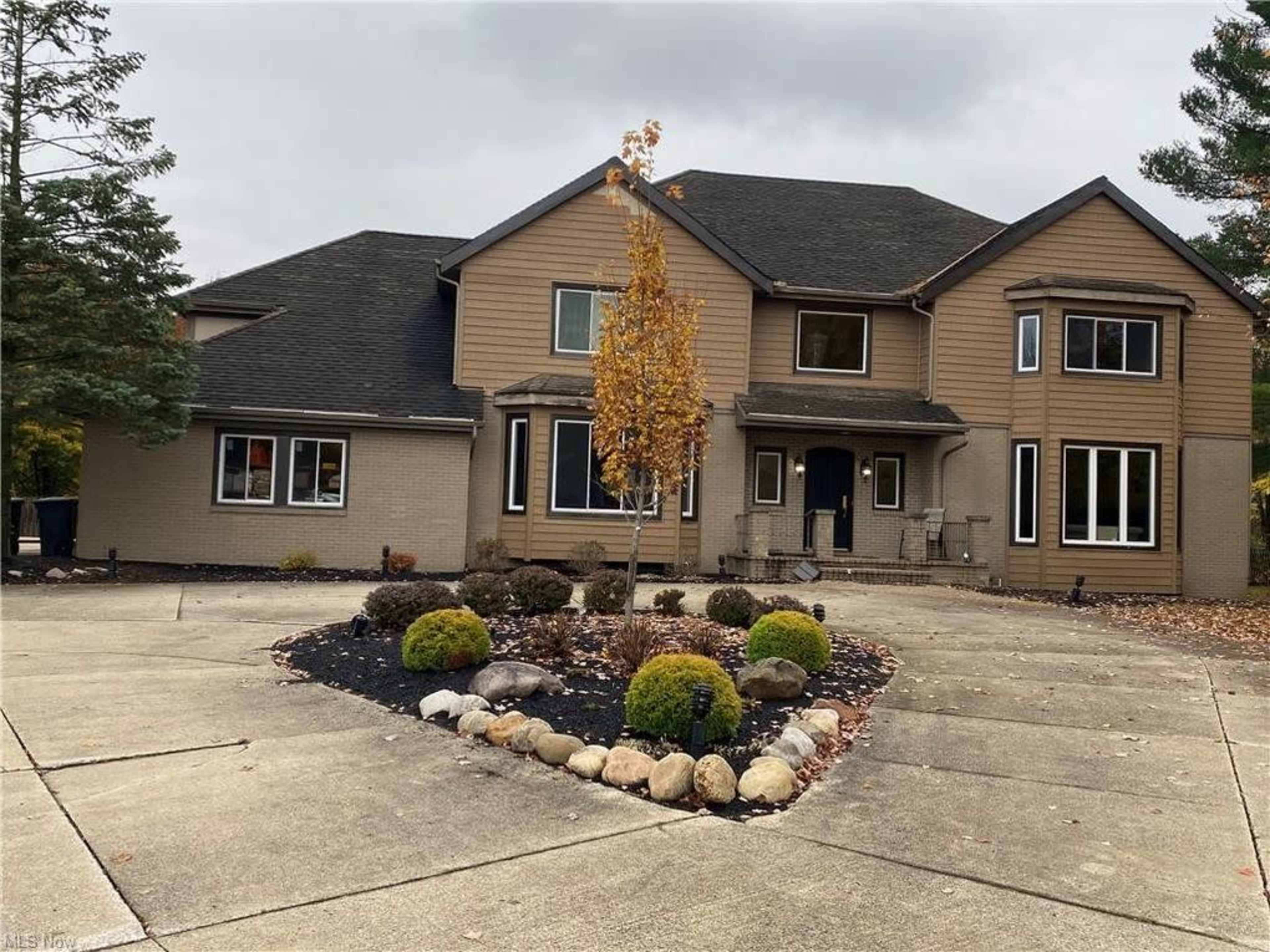 A large, two-story house with a brown and beige exterior, featuring landscaped grounds and a circular driveway.