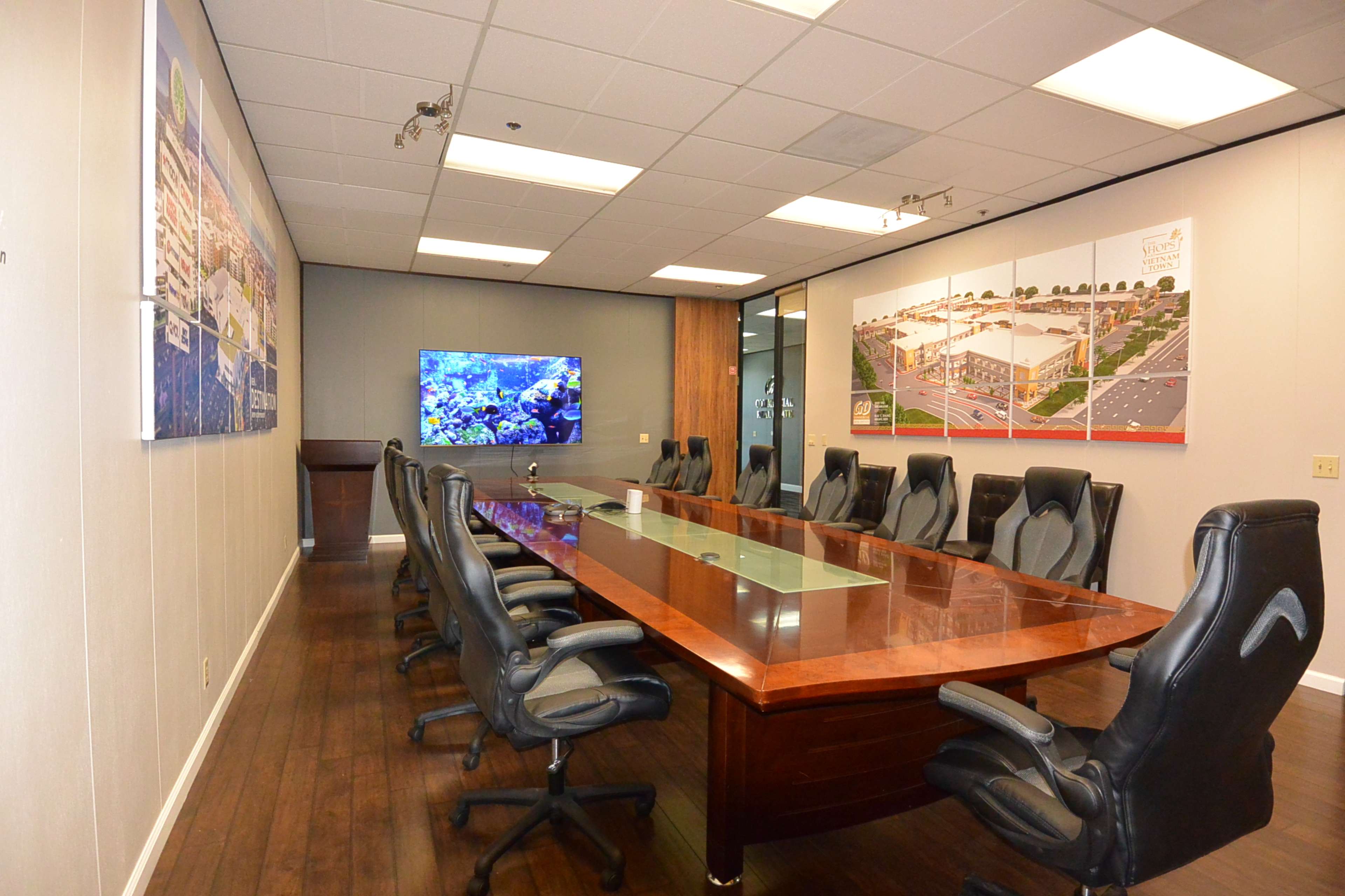 A large conference room features a long wooden table surrounded by black leather chairs, with a television and informational displays on the walls.