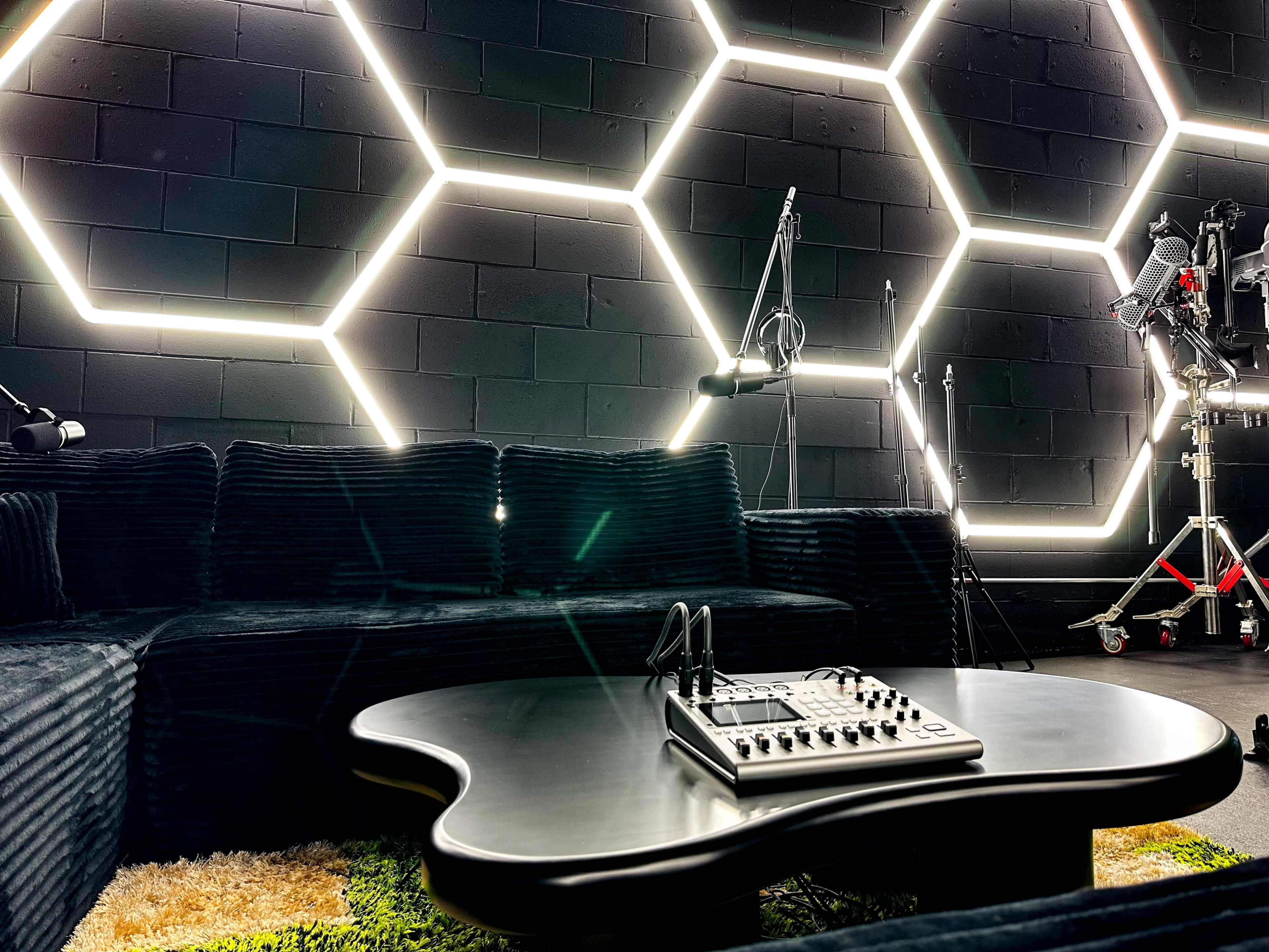 A modern lounge area features a black hexagon-patterned wall illuminated by neon lights, with a low, irregularly shaped coffee table and audio equipment set on a textured carpet.