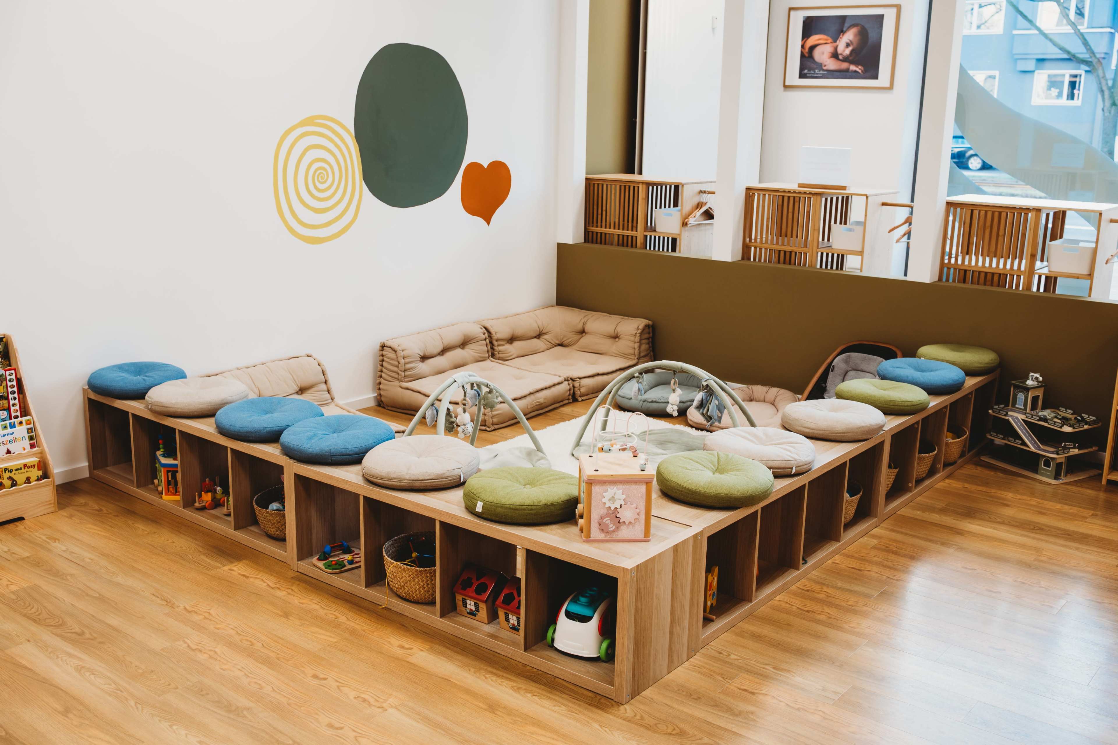 The image shows a play area with a circular seating arrangement made of cushions and low wooden storage units filled with toys and baskets.