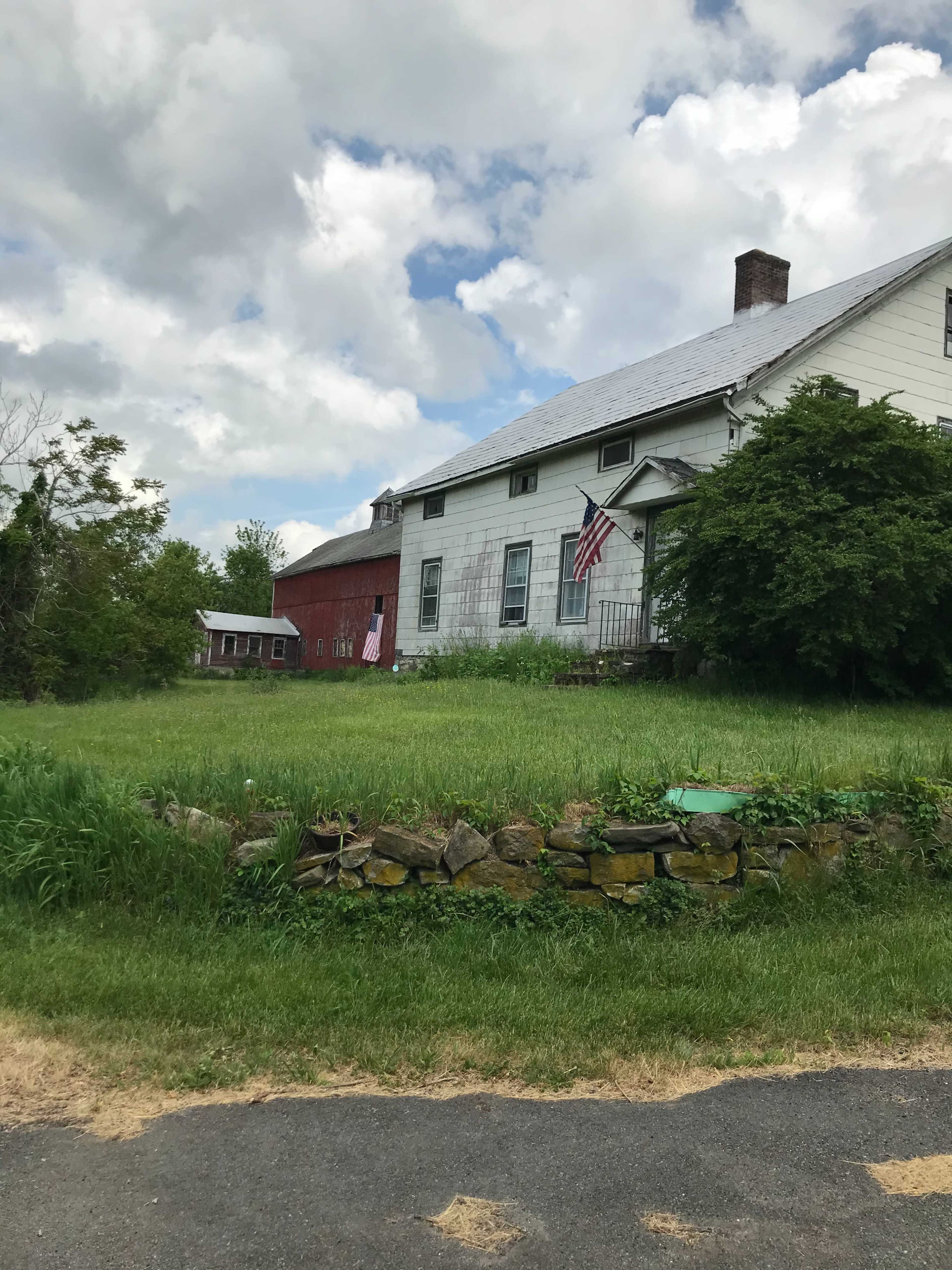 A white farmhouse with a gray roof and American flags hangs on the front, bordered by green grass and a stone wall, next to a red barn in a rural setting.