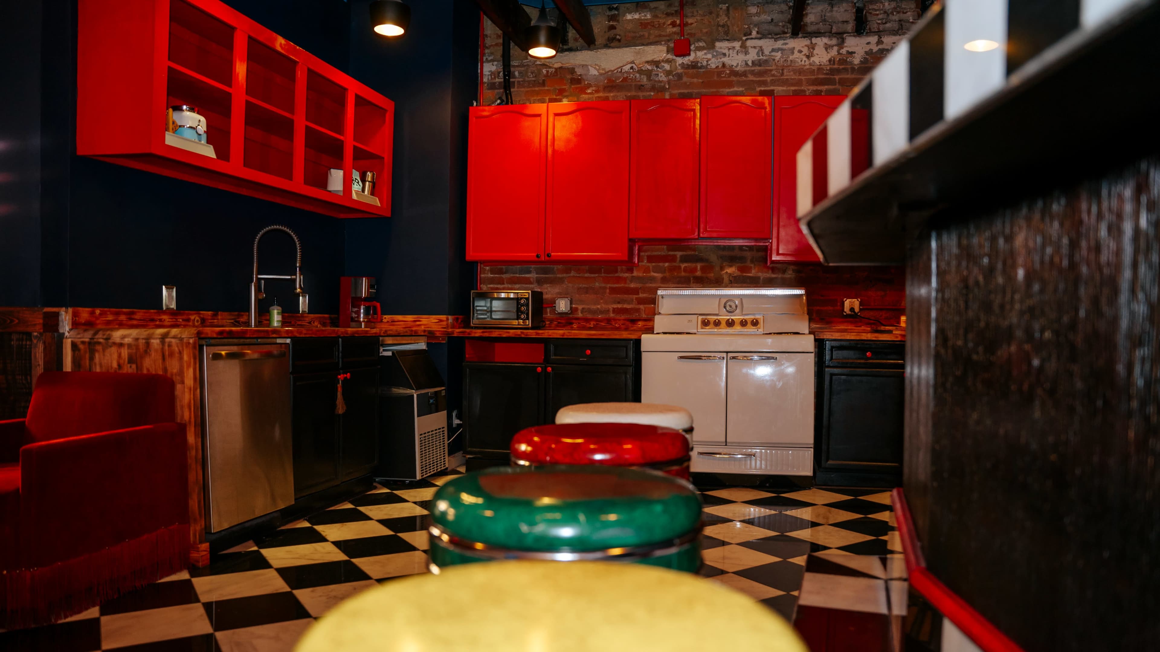 The image shows a kitchen with a checkered black and white floor, stainless steel appliances, and colorful bar stools.