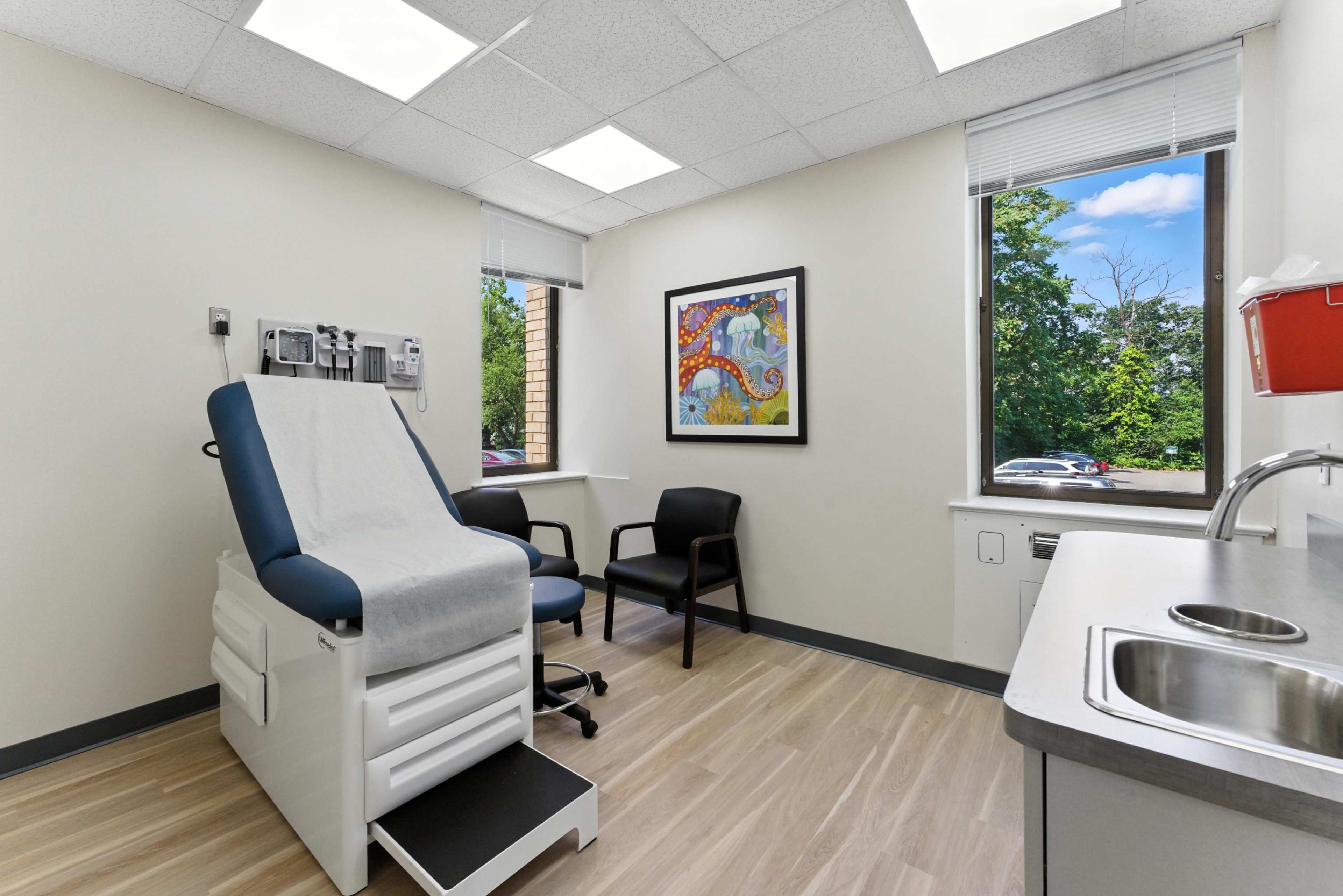 The image shows a doctor's examination room with a medical examination table, two chairs, a window with a view of trees, and a small sink area.