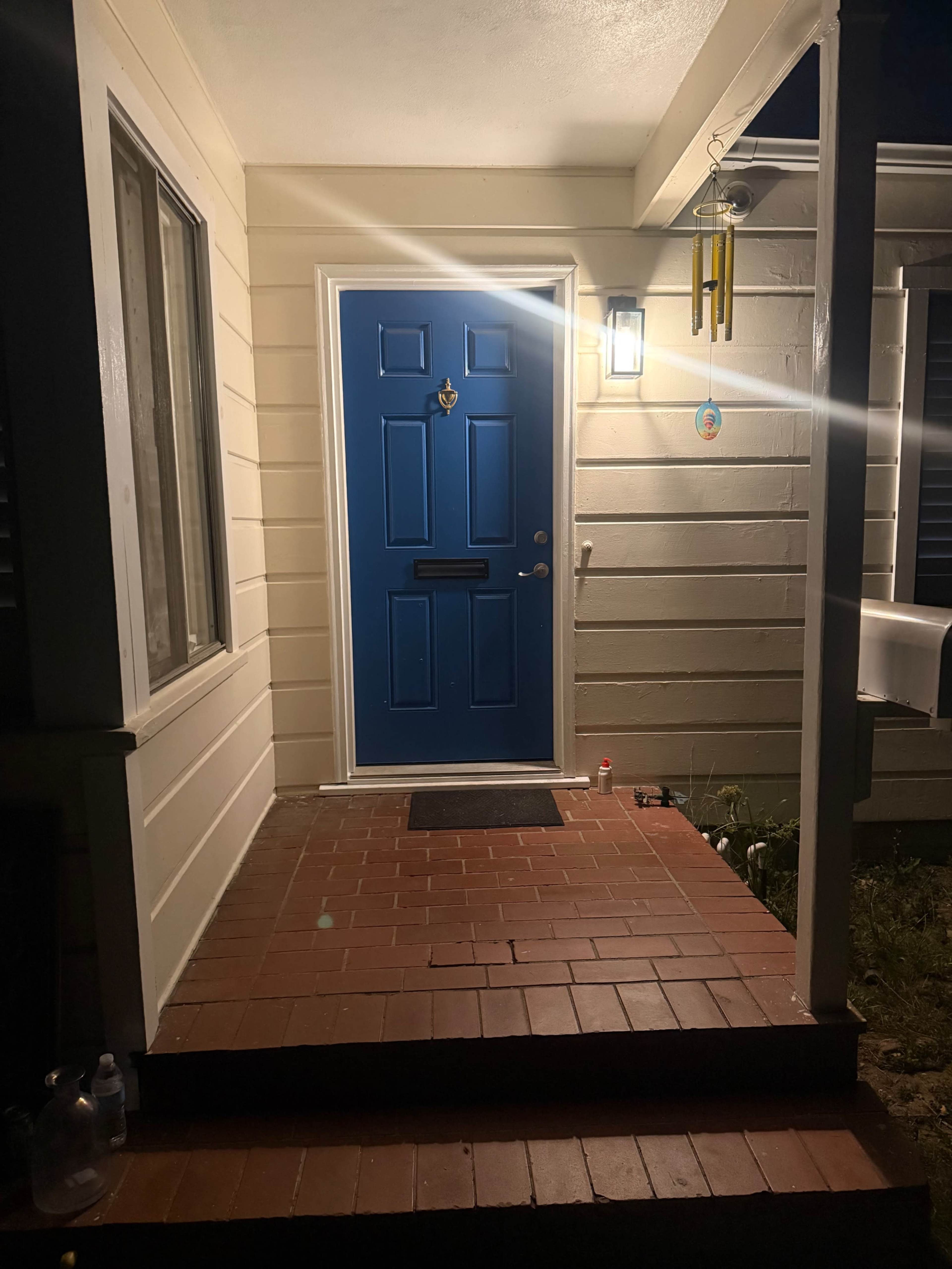 The image shows a house entrance with a blue front door, a light fixture, a welcome mat, and a small wind chime hanging nearby.