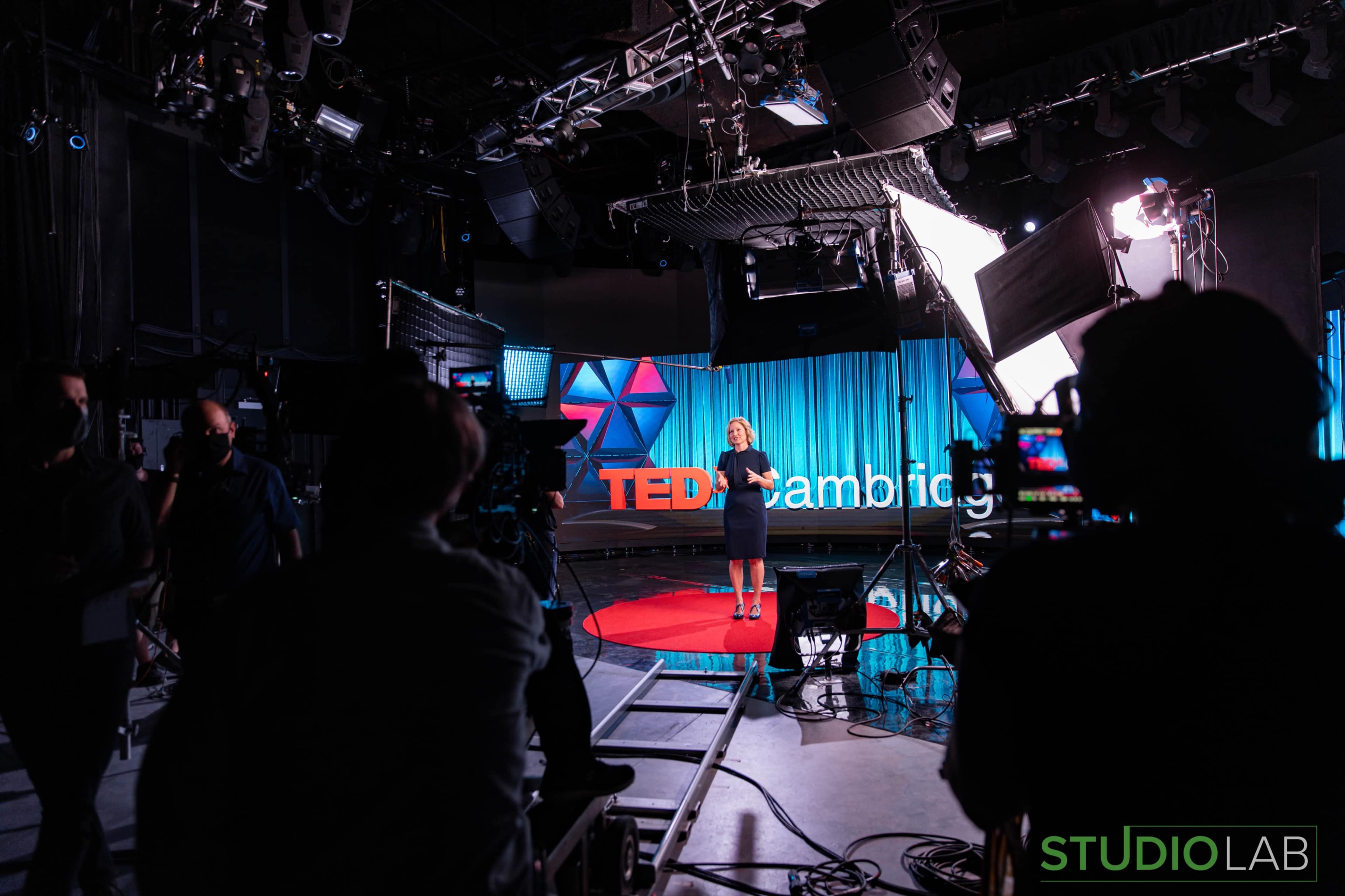 A speaker stands on stage at a TED event, with cameras and lighting equipment set up in the studio.