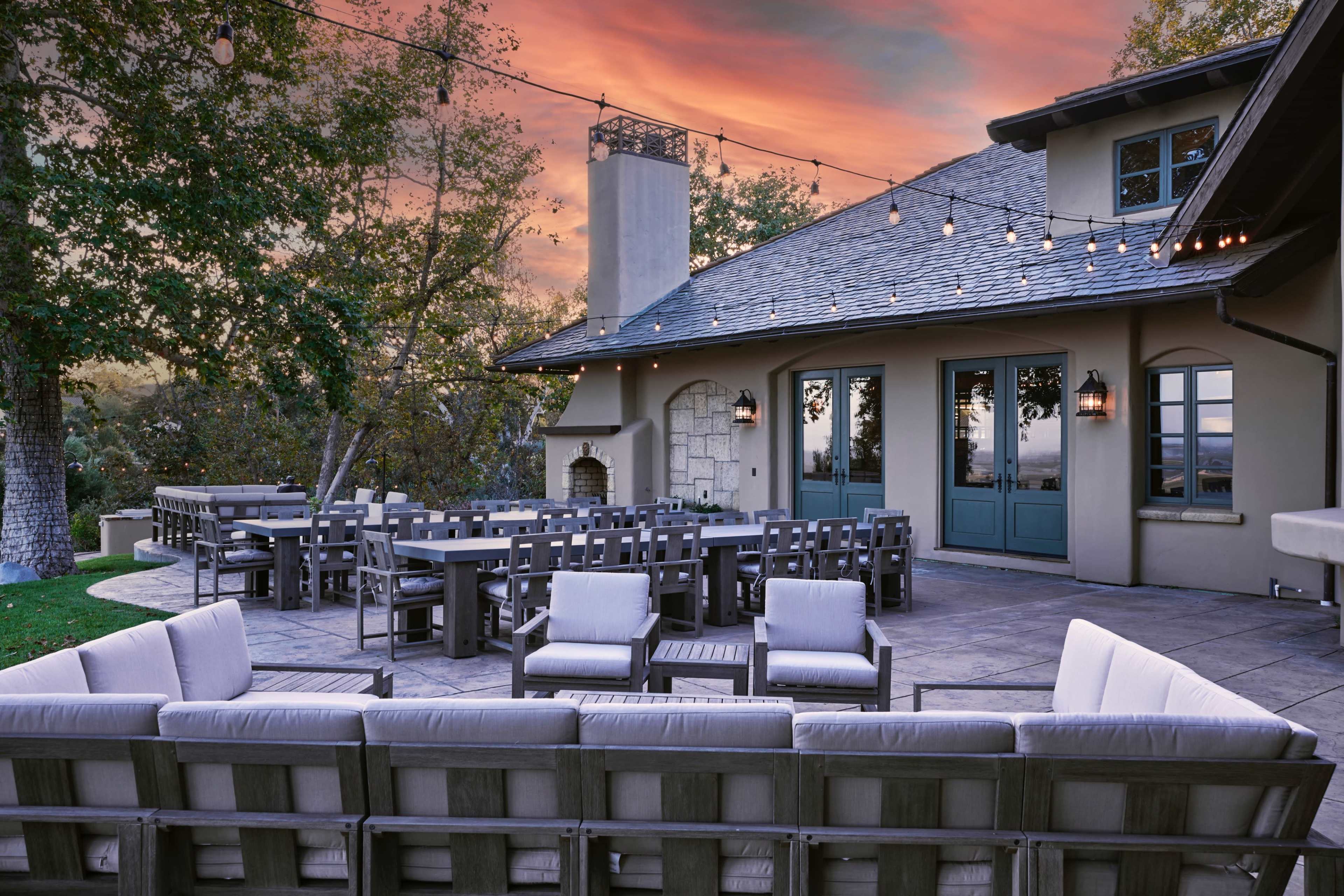 The scene features a patio with outdoor seating arranged in a circular layout, adjacent to a house with large windows and a stone fireplace, under a sky painted with sunset colors.