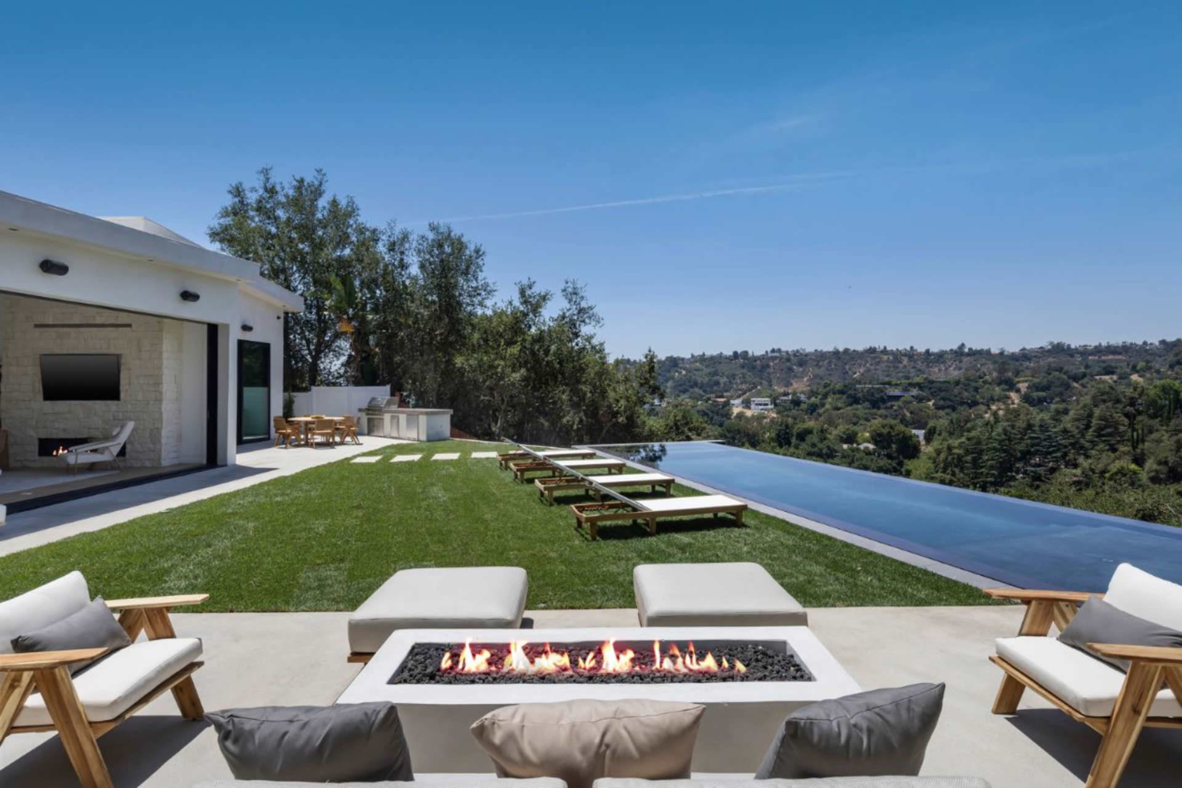 The image features a modern outdoor living space with a fire pit, lounge chairs, and an infinity pool overlooking a lush landscape.