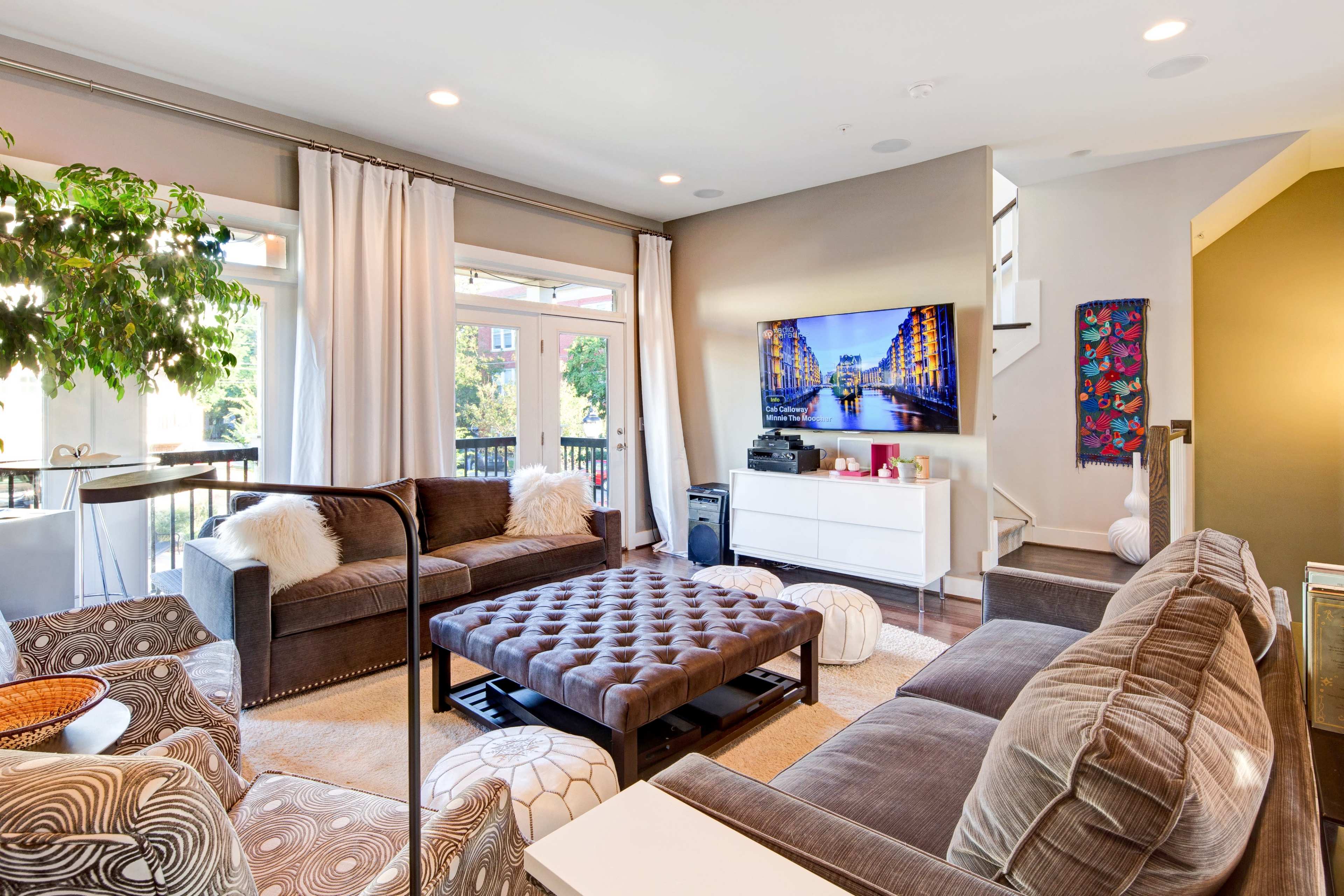 The living room features a large sectional sofa, a coffee table, and a television mounted on the wall, illuminated by natural light from large windows.