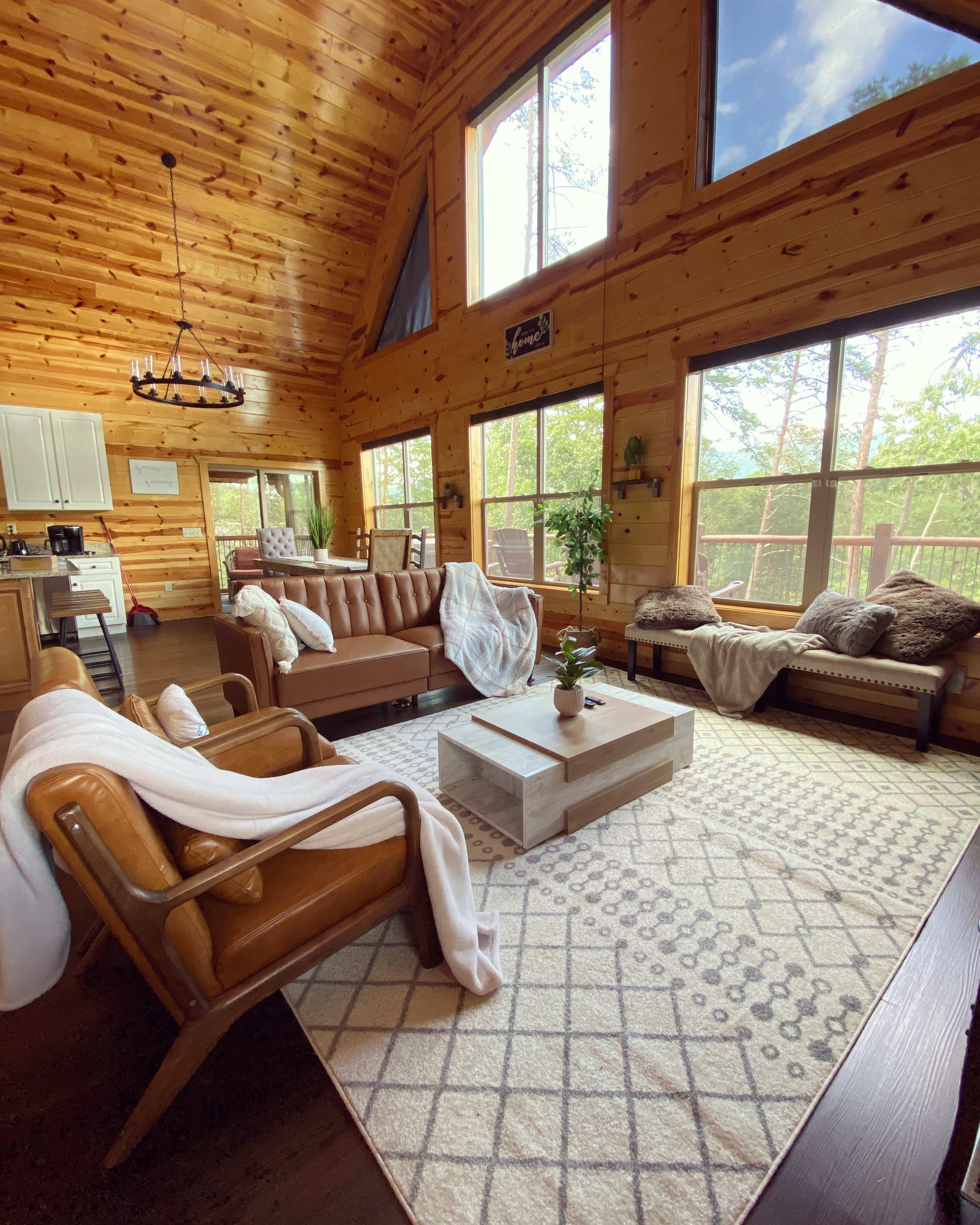 The image shows a cozy living room with wooden walls, large windows, and a mix of seating furniture, including a sofa and armchairs, around a central coffee table on a patterned rug.
