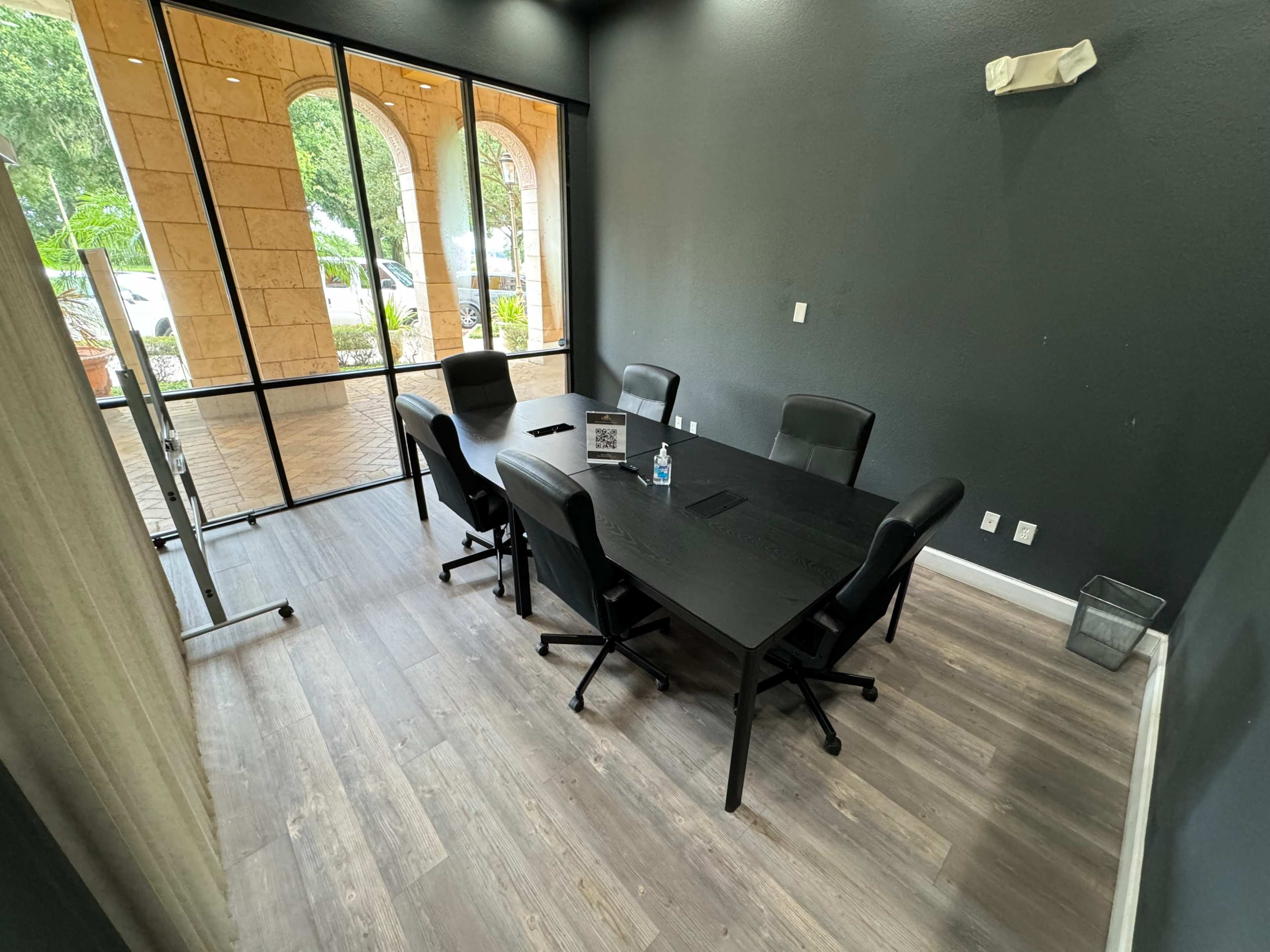 A modern conference room features a large table surrounded by six black chairs and a glass wall showcasing views of the outside area.