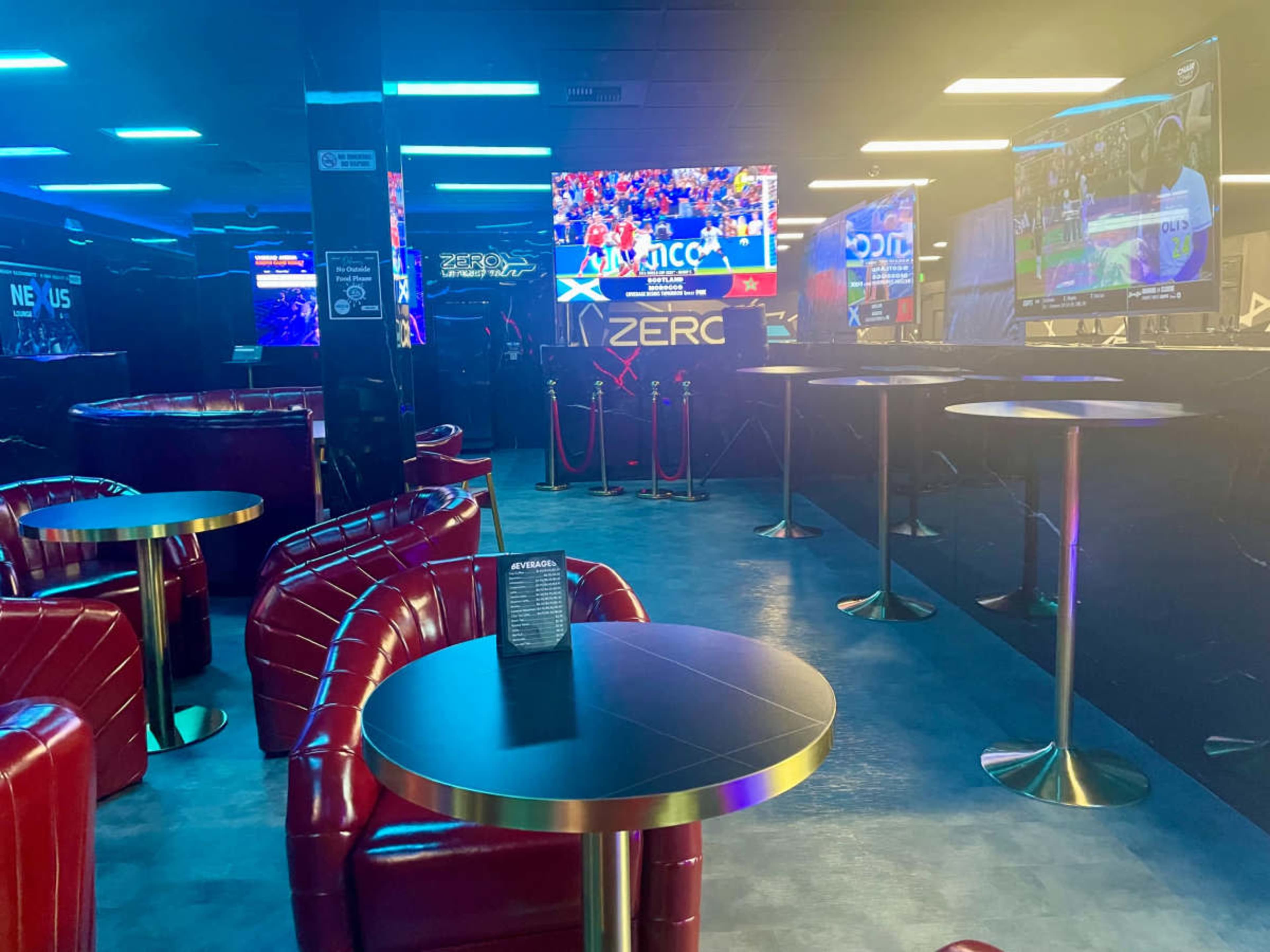 The image shows a modern lounge area with red leather seating and several small tables, featuring multiple screens displaying sports events in a dimly lit environment.