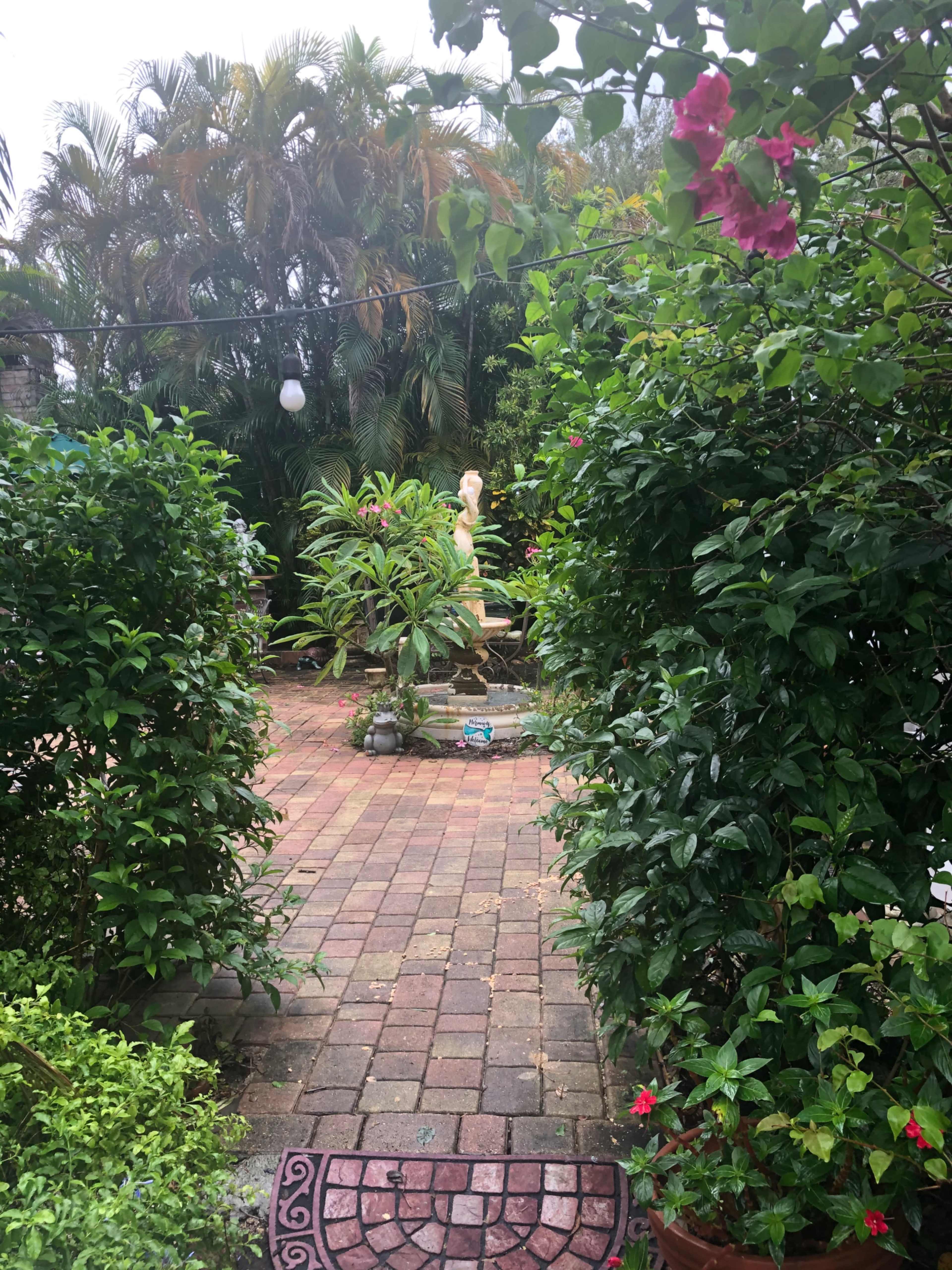 The image shows a garden pathway lined with greenery leading to a statue in a brick-paved area.