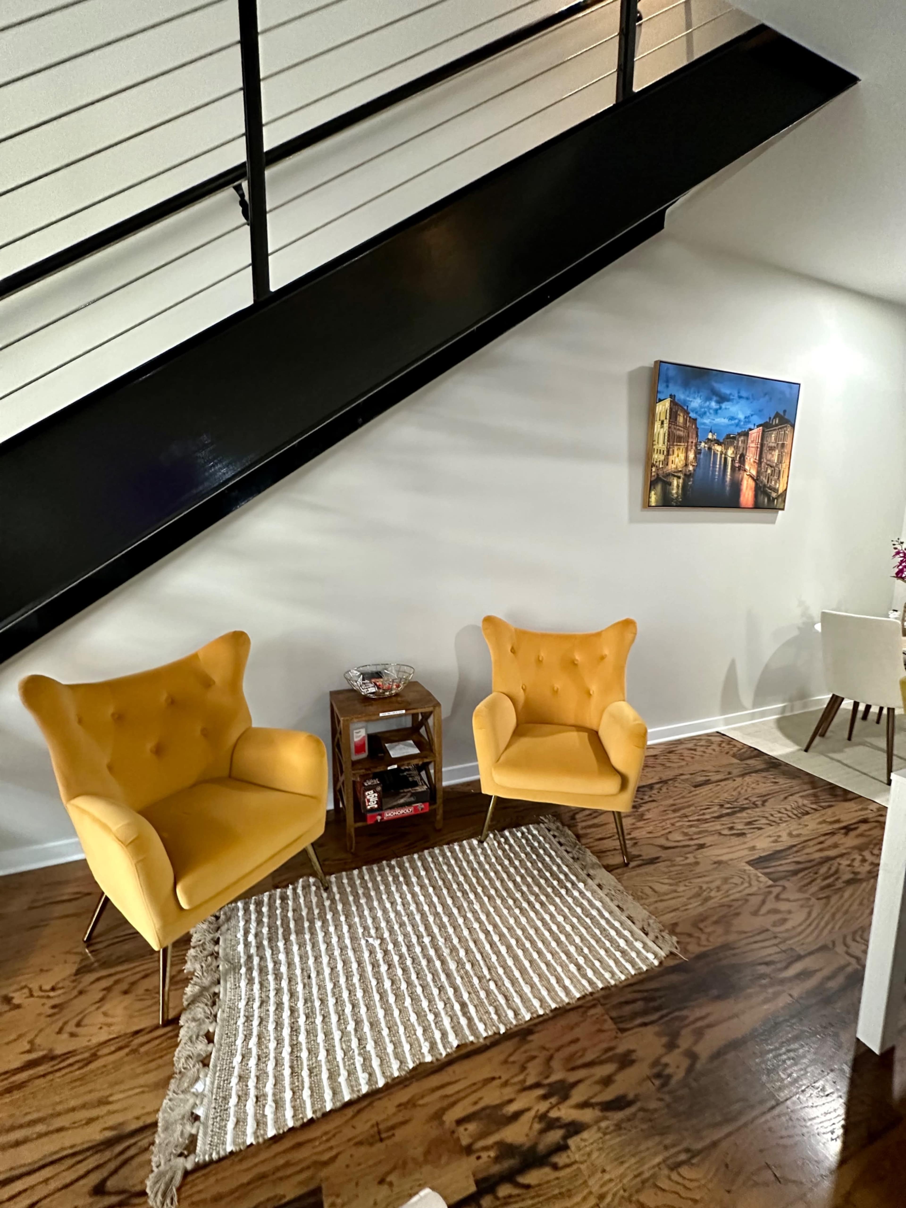 Two yellow armchairs are positioned on a rug in a living area beneath a staircase, with a small table nearby and a framed picture on the wall.