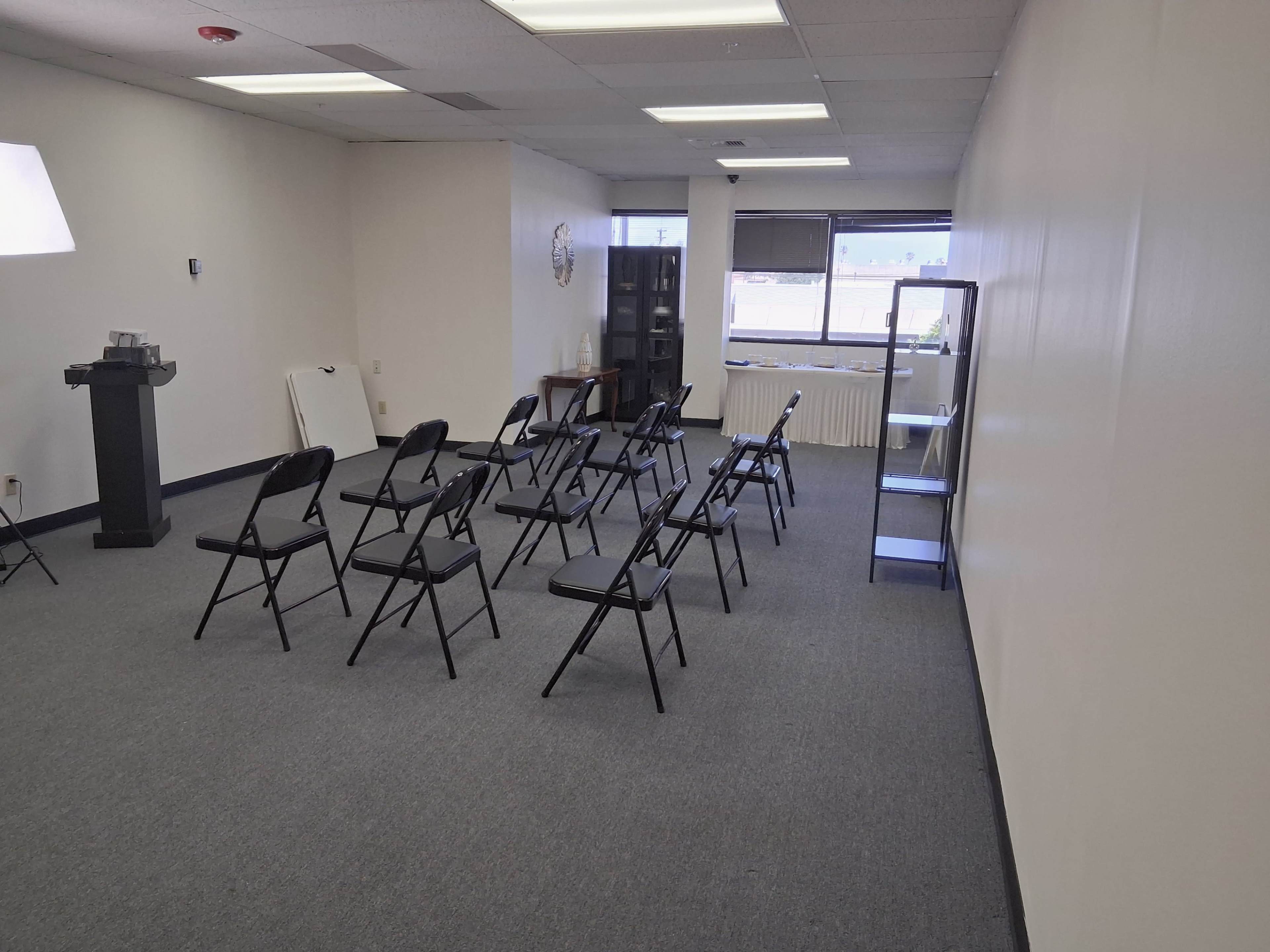 The image shows a sparsely furnished room with several black folding chairs arranged in rows and a few display shelves against the walls.