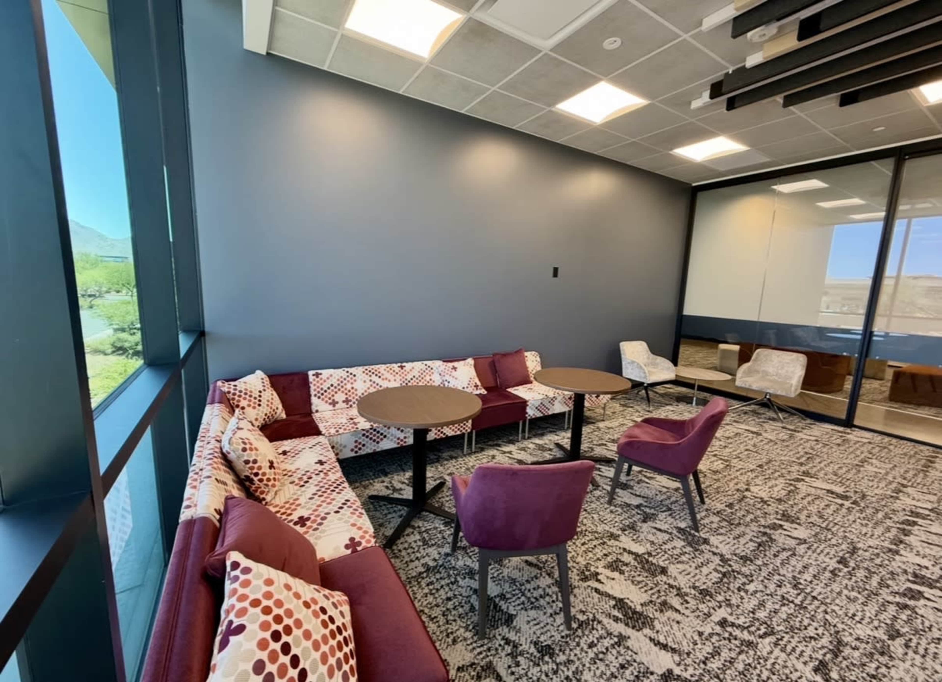 A modern lounge area features a maroon and white patterned couch, two round tables, and several chairs near large windows.