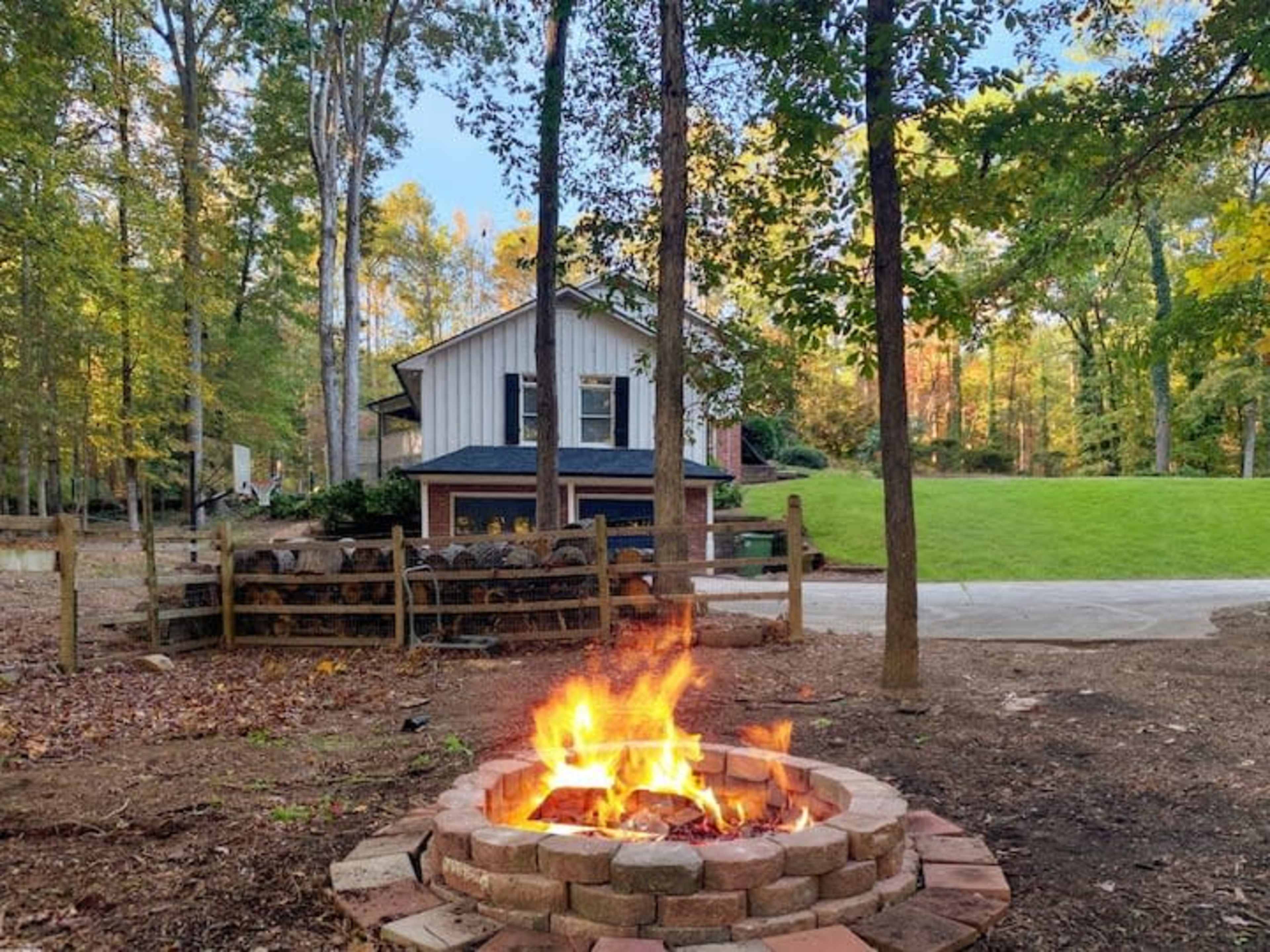 A stone fire pit with a vibrant fire is surrounded by trees, facing a two-story house in a wooded area.