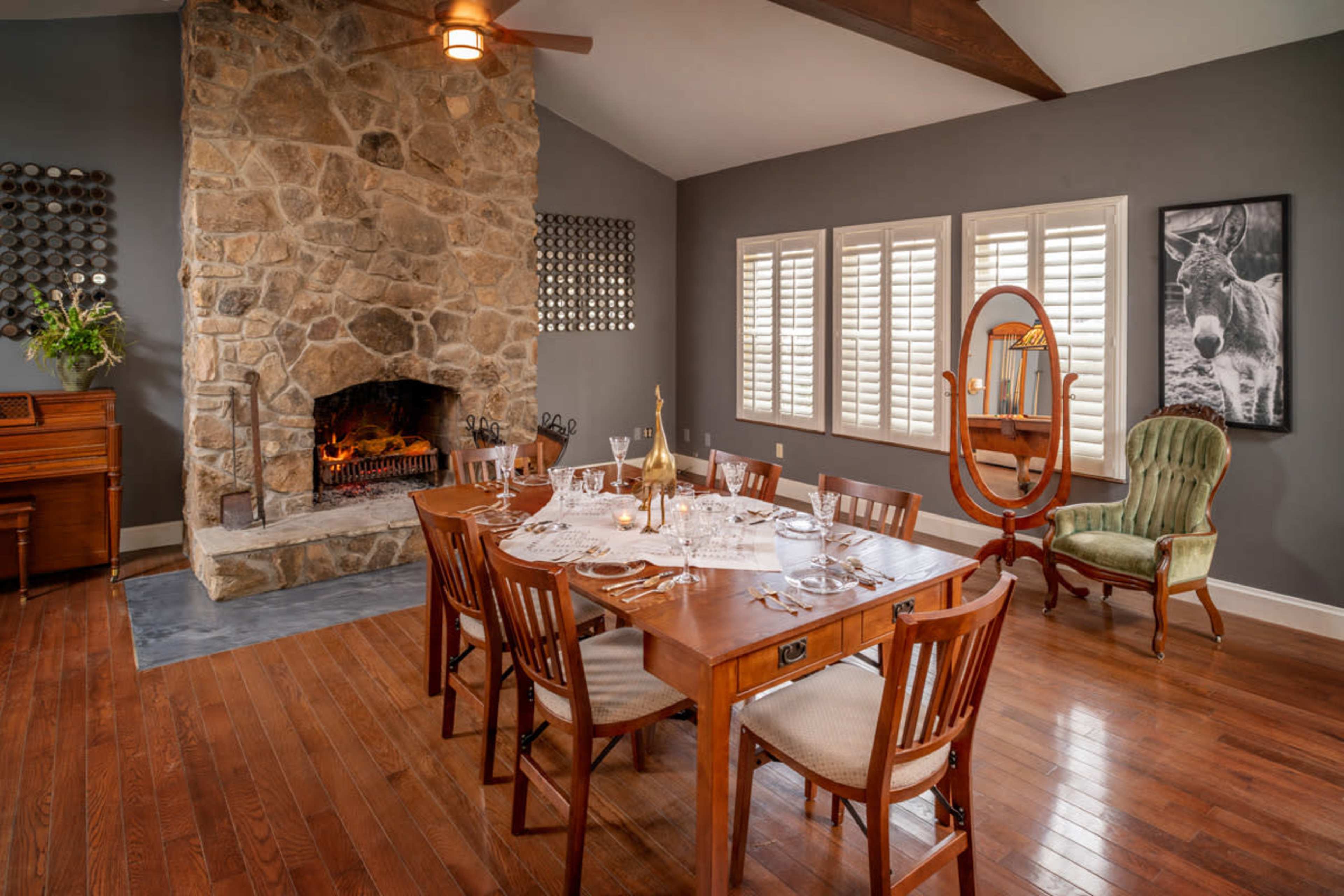 A wooden dining table is set with dishes and utensils in a room featuring a stone fireplace and large windows with shutters.
