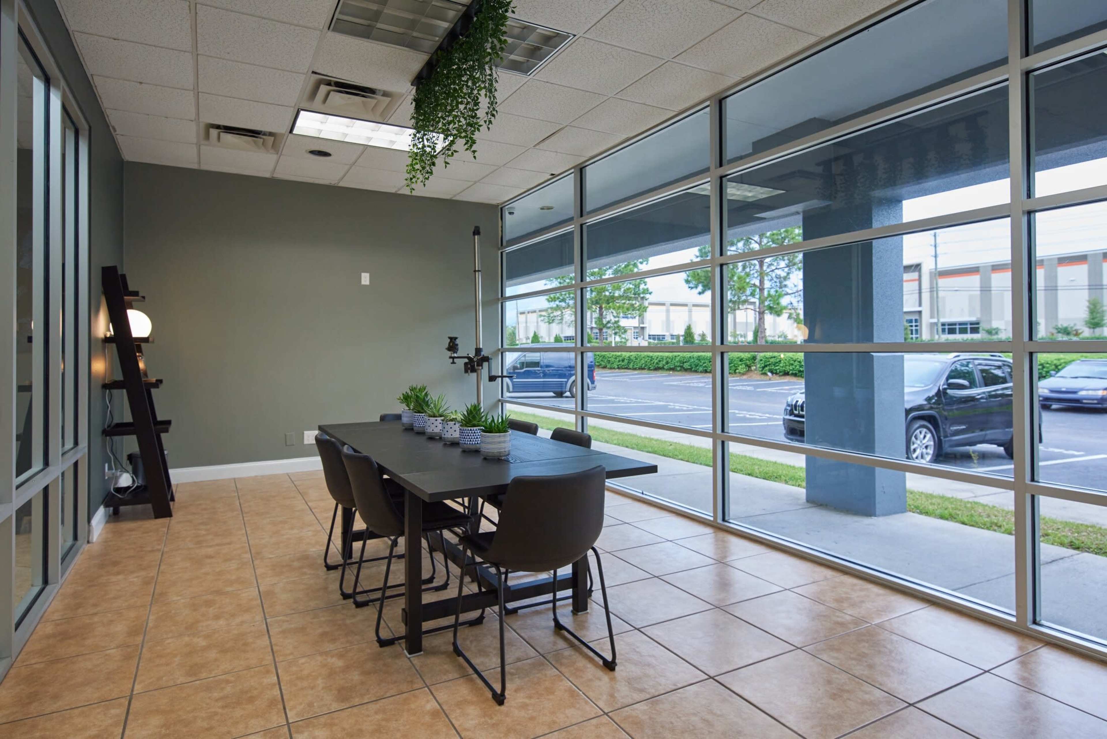 A modern meeting room features a large table with chairs, surrounded by windows offering a view of an outdoor parking area.