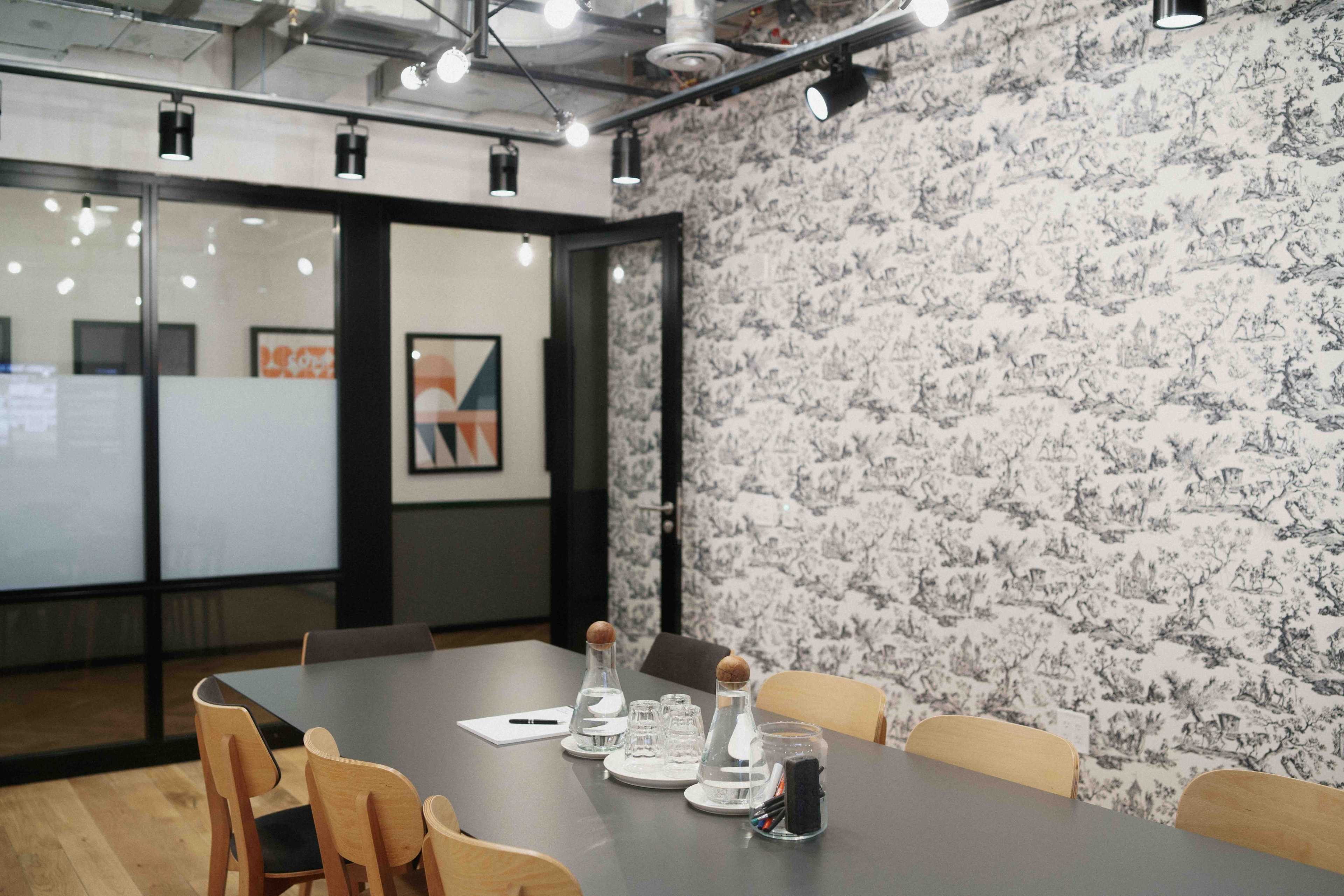 A modern meeting room with a long table set with glass water pitchers, cups, and a notepad, surrounded by patterned wallpaper and track lighting.