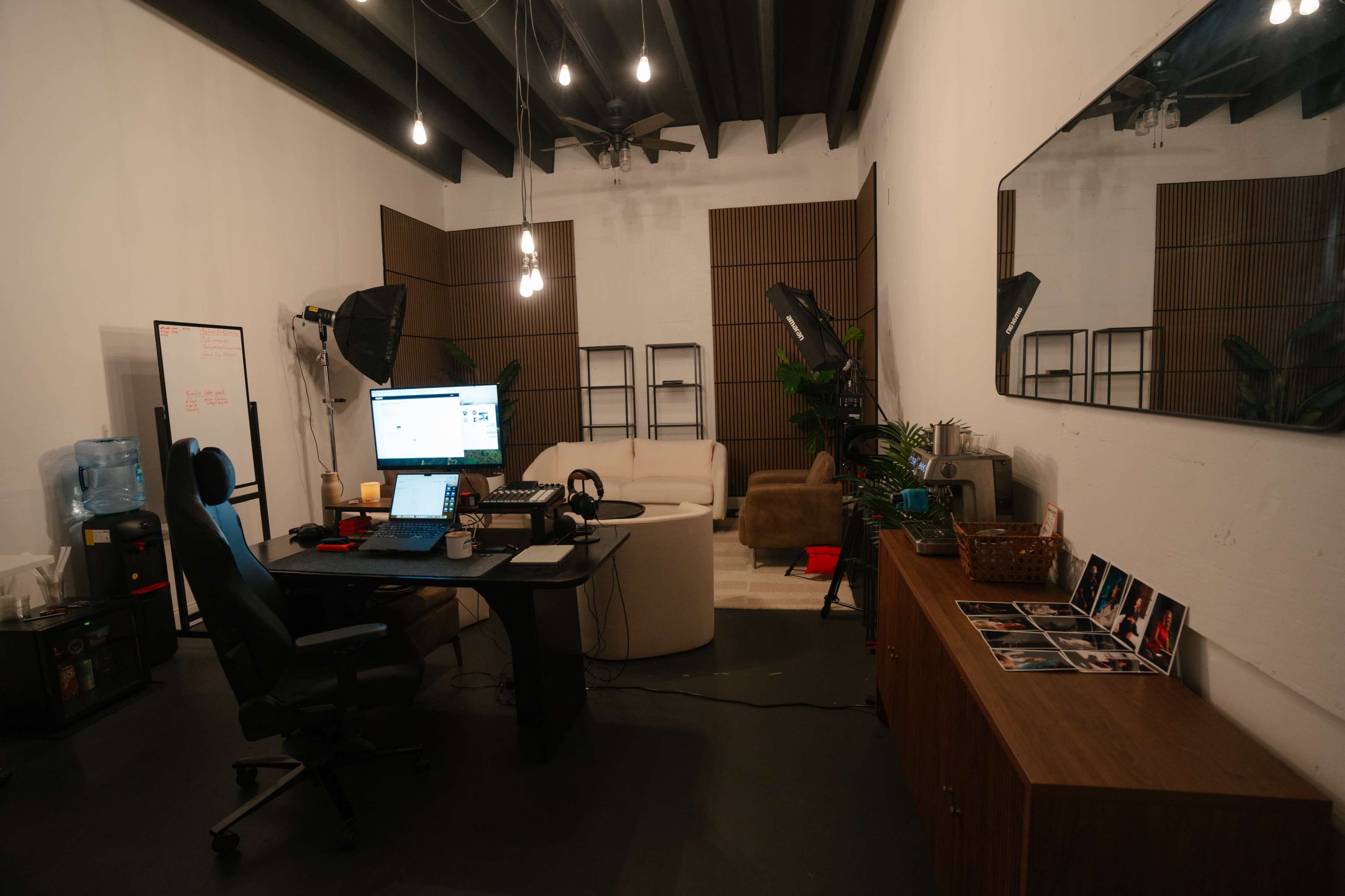 The image shows a modern office space featuring a desk with a computer setup, a whiteboard, a couch, plants, and various light fixtures hanging from the ceiling.