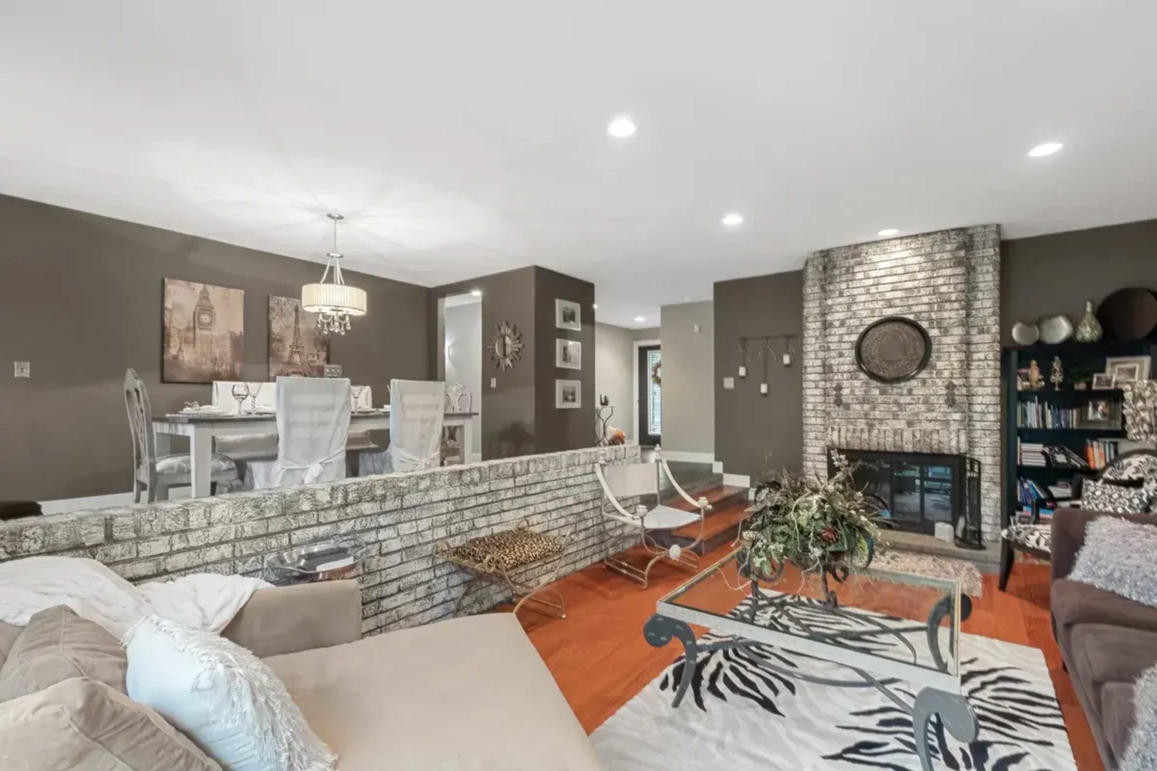 A living room with a stone fireplace, hardwood flooring, and a modern dining area visible in the background.