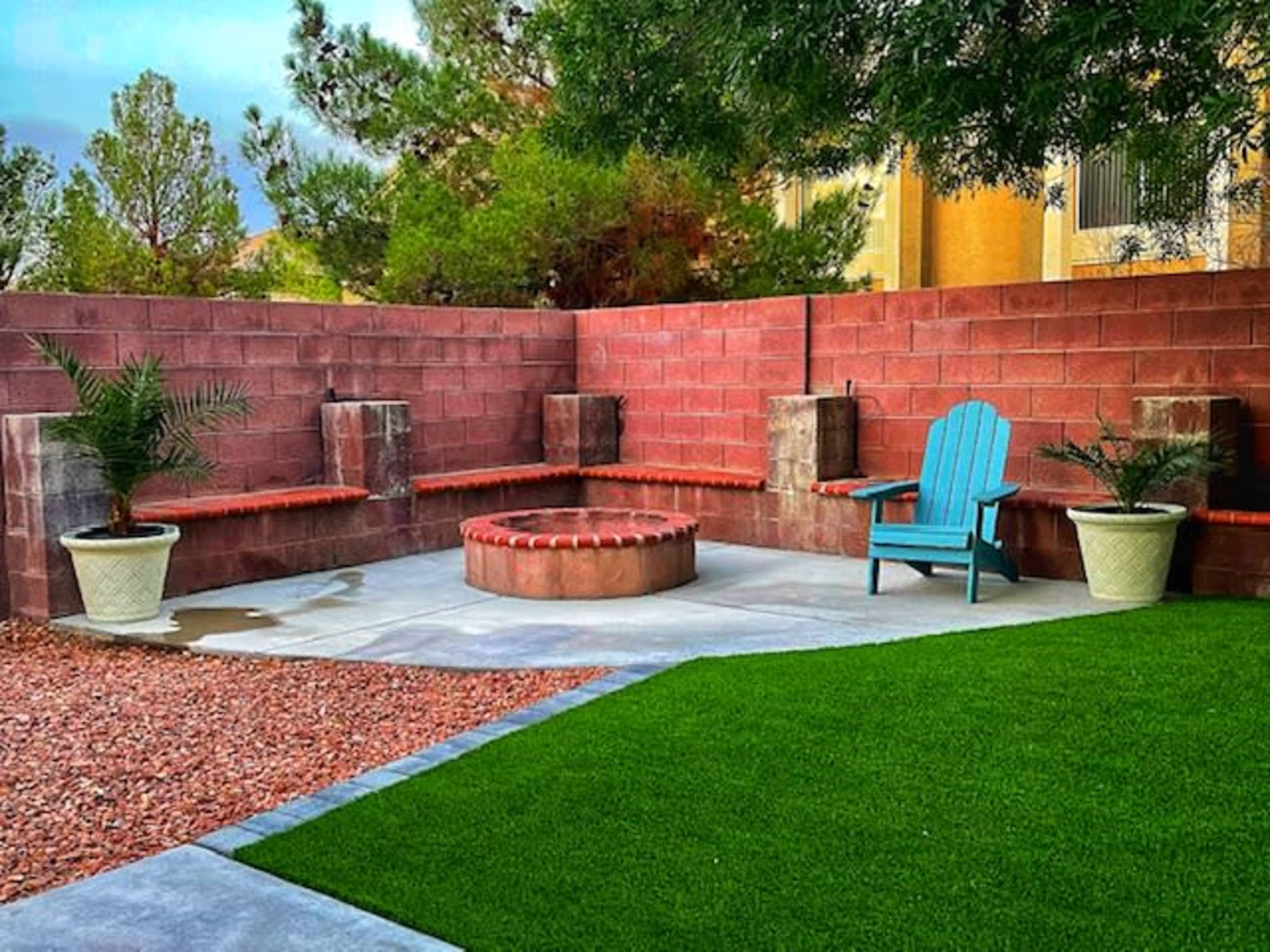 The image shows a backyard area featuring a circular fire pit surrounded by two benches, a blue adirondack chair, and potted plants on either side, set against a wall with a well-kept lawn adjacent to gravel.