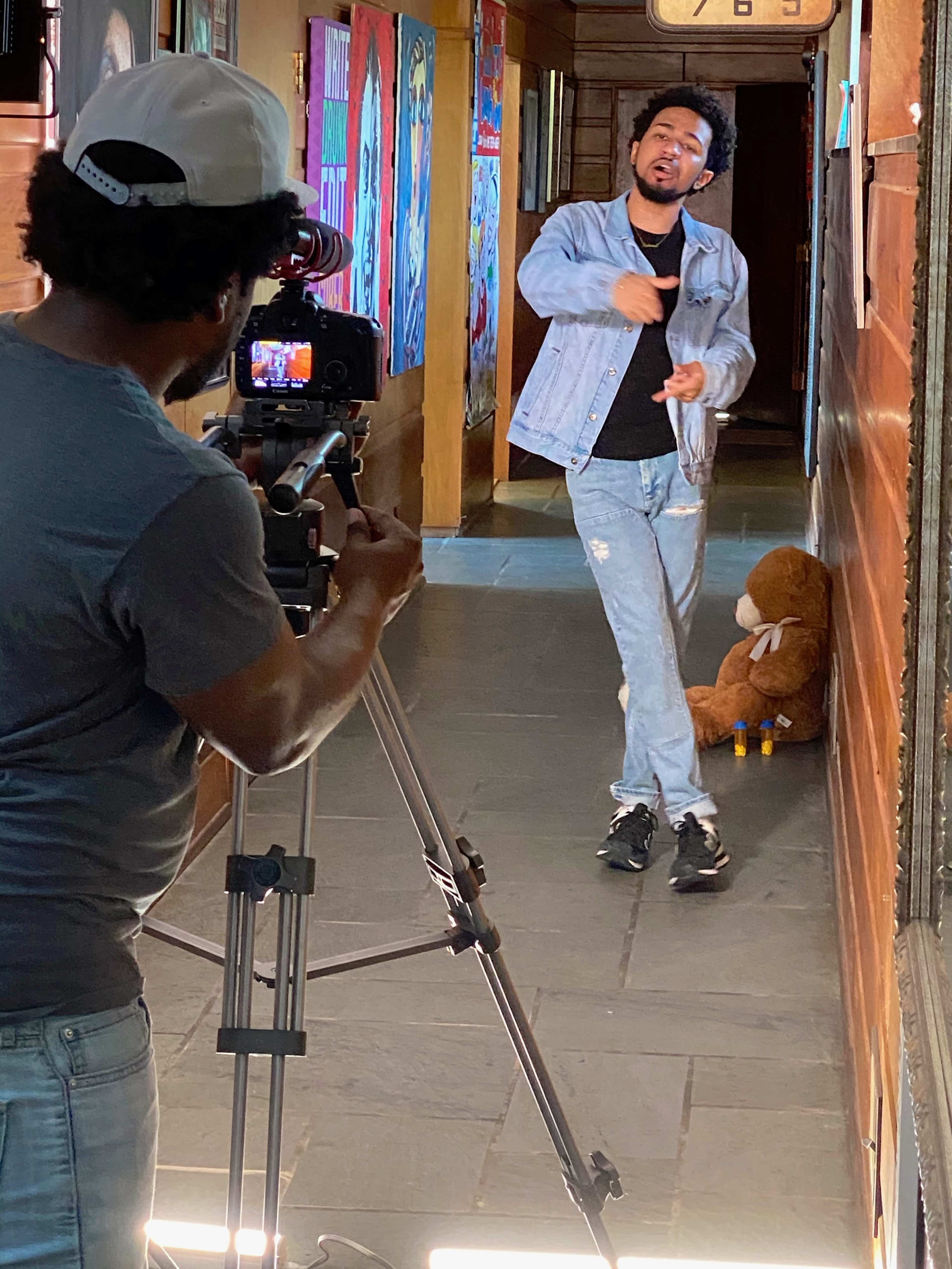 A person wearing a denim jacket performs in a hallway while another individual records the scene with a camera set on a tripod.