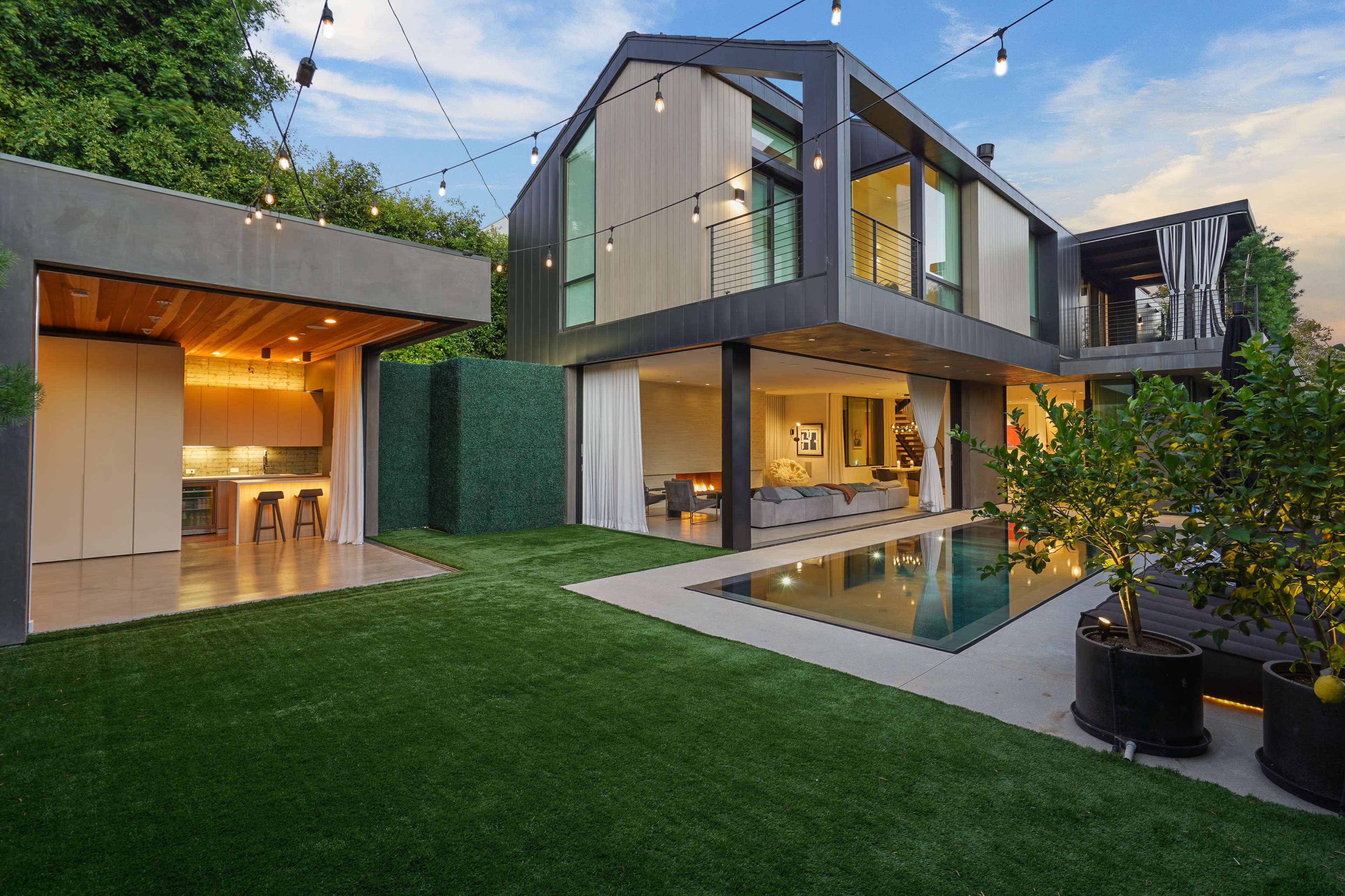 A modern house with a swimming pool and landscaped yard features large glass windows and outdoor string lights.