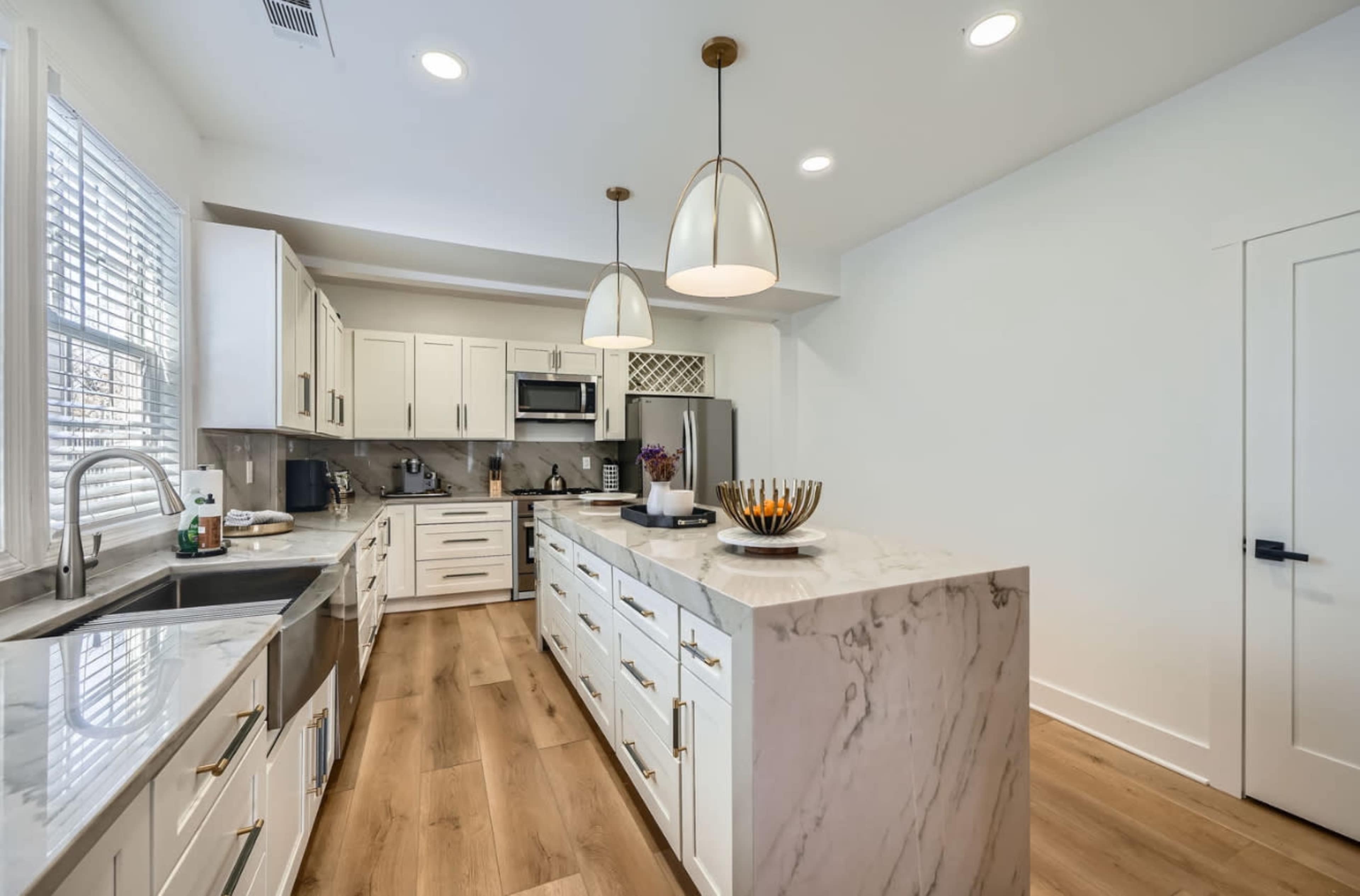 The image shows a modern kitchen with white cabinetry, a marble countertop, stainless steel appliances, and pendant lighting.
