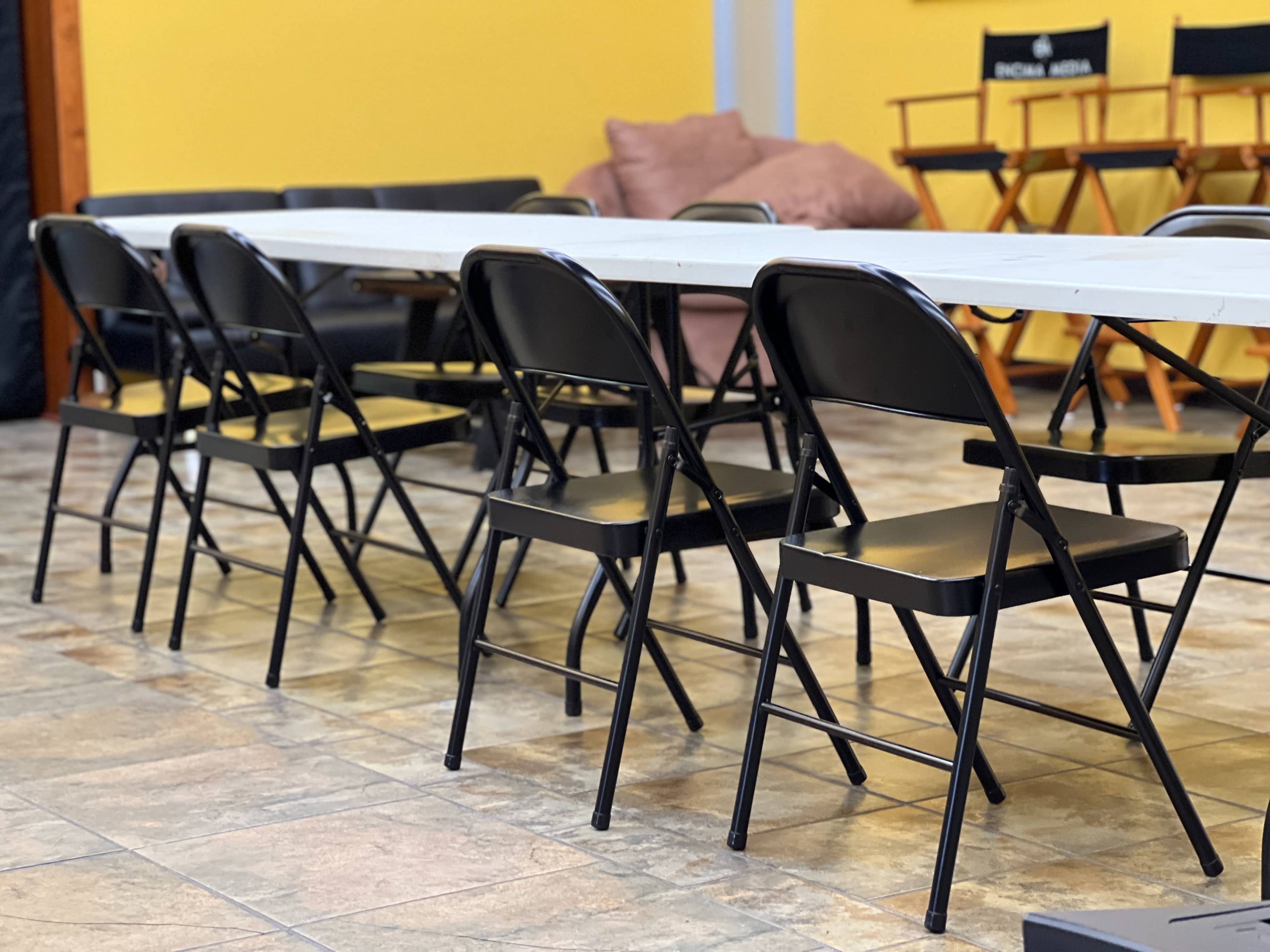 A row of black folding chairs is positioned beside a long white table in a room with yellow walls.
