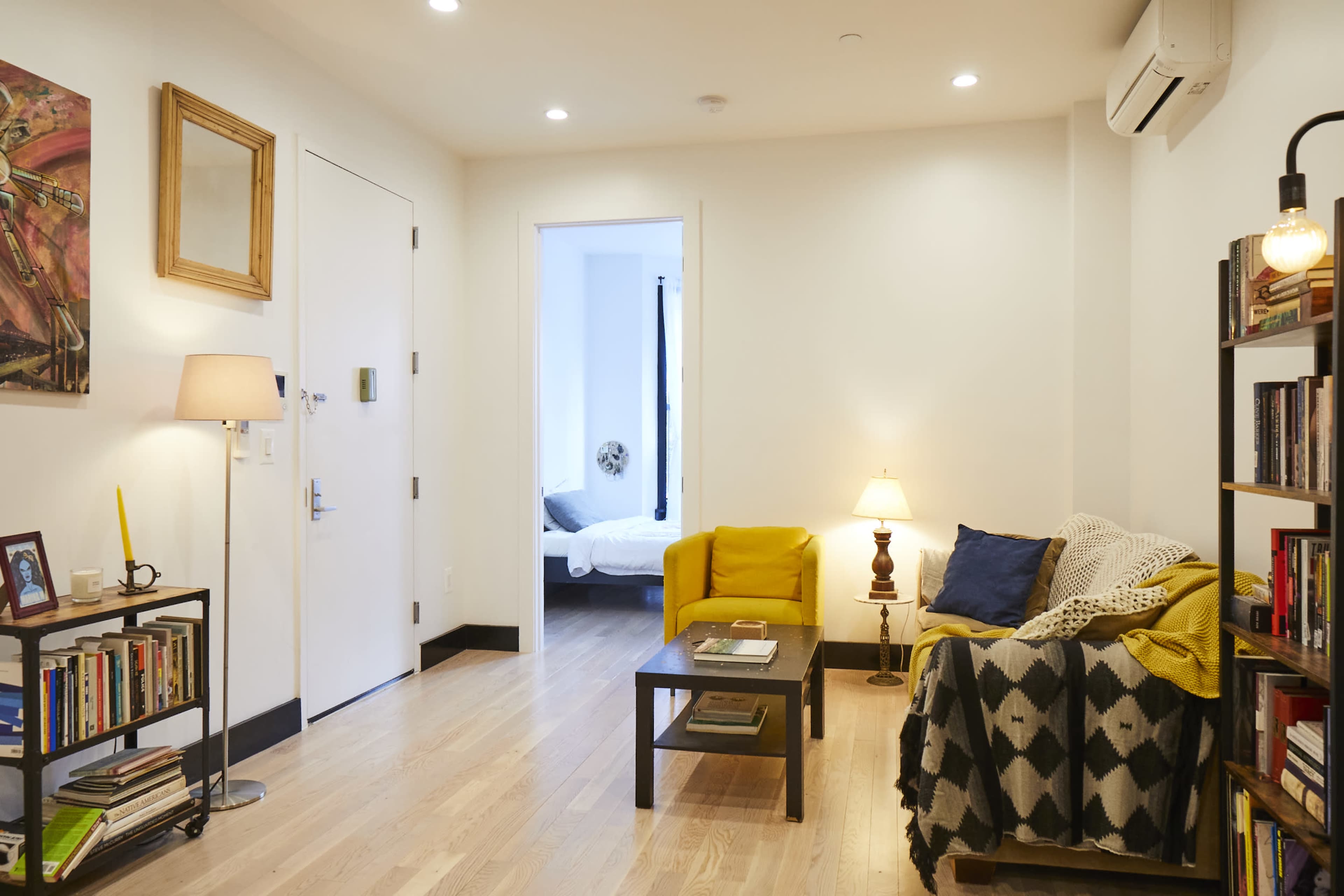 The image depicts a modern living room with yellow seating, a black bookshelf filled with books, and a doorway leading to a bedroom.