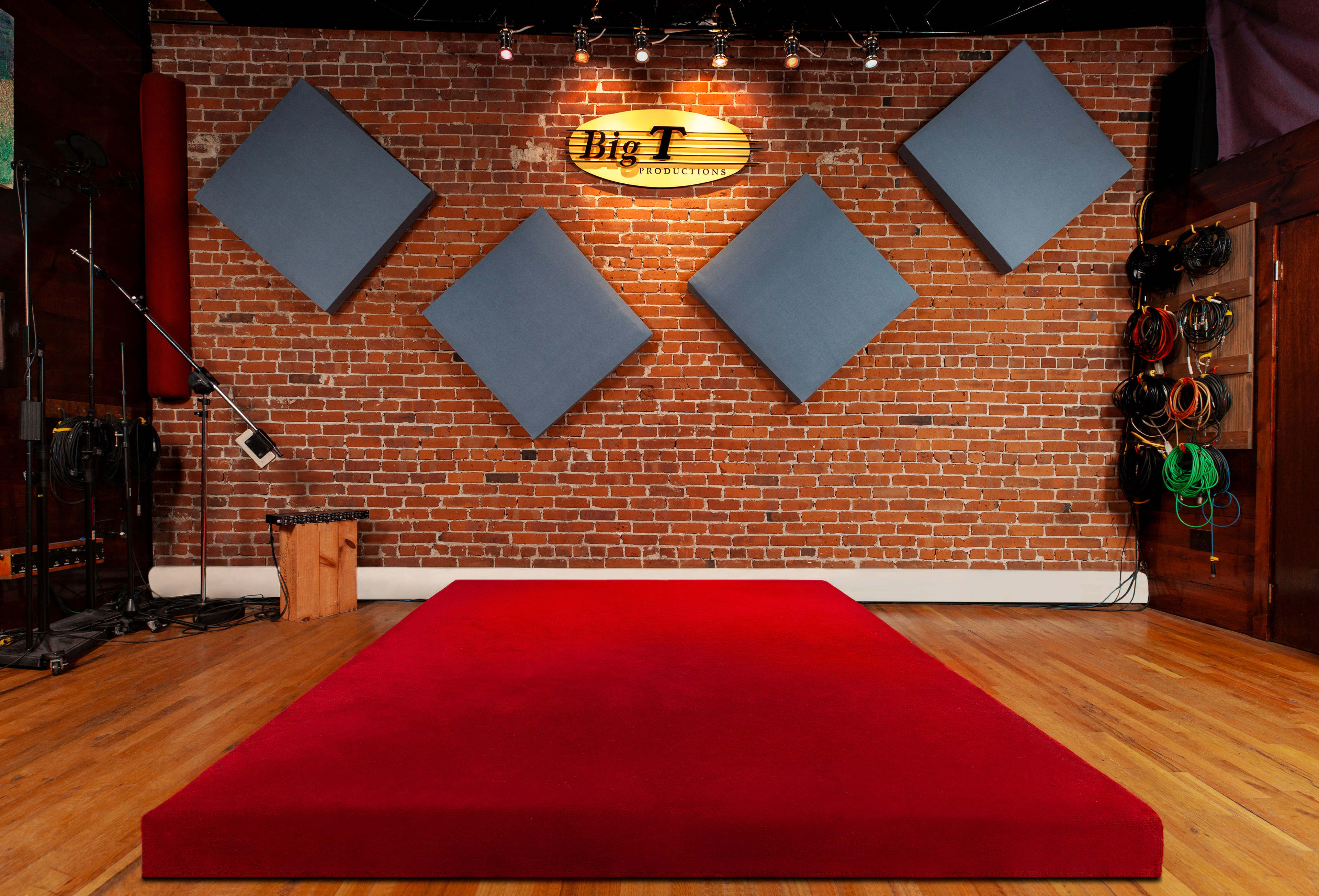 A stage area with a red carpet in front of a brick wall featuring four blue sound-absorbing panels and a sign that reads "Big T Productions."