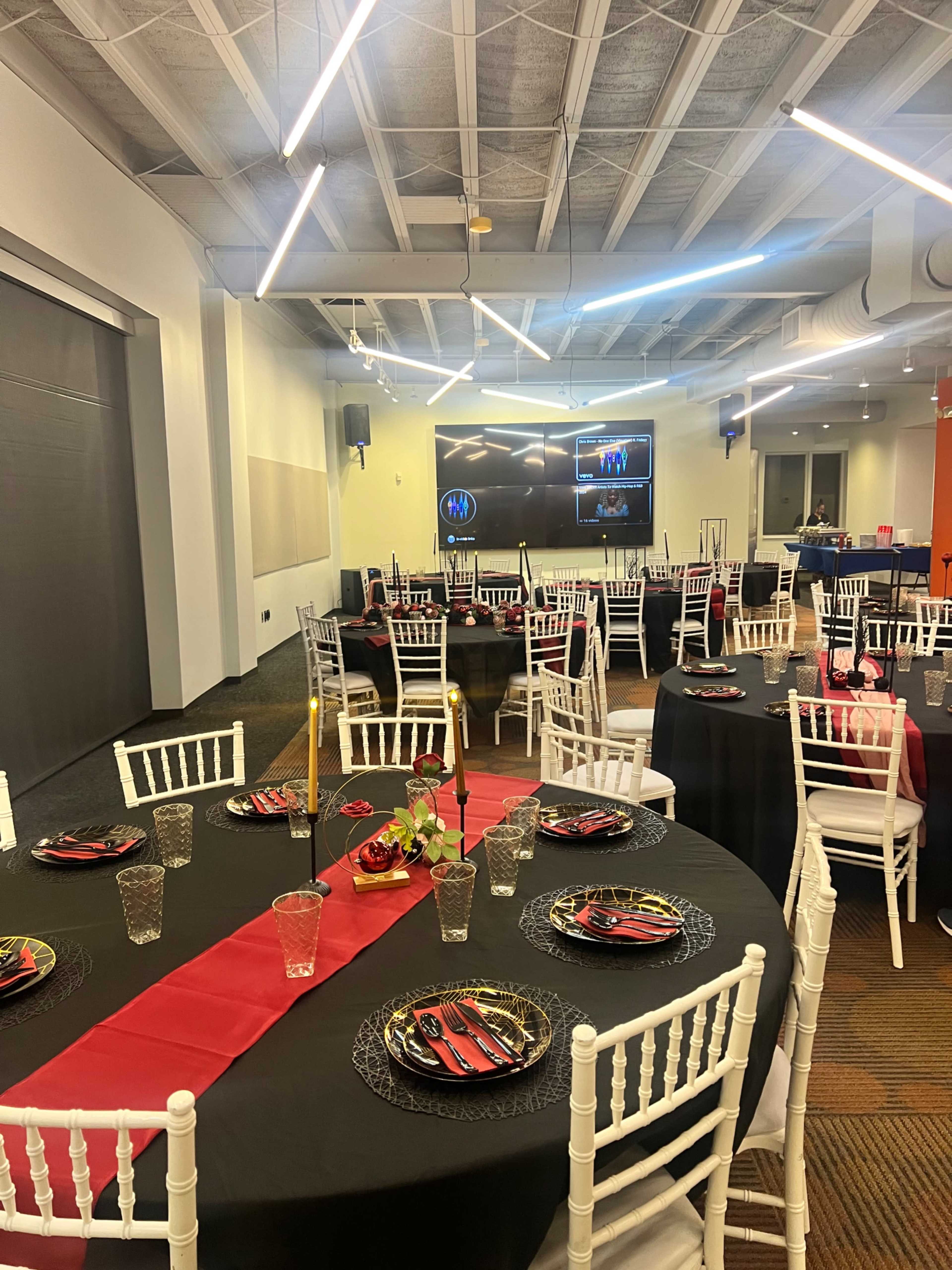 A banquet hall is set up with tables covered in black and red tablecloths, adorned with plates and glasses, and chairs placed around them, while a screen displays images at the front of the room.