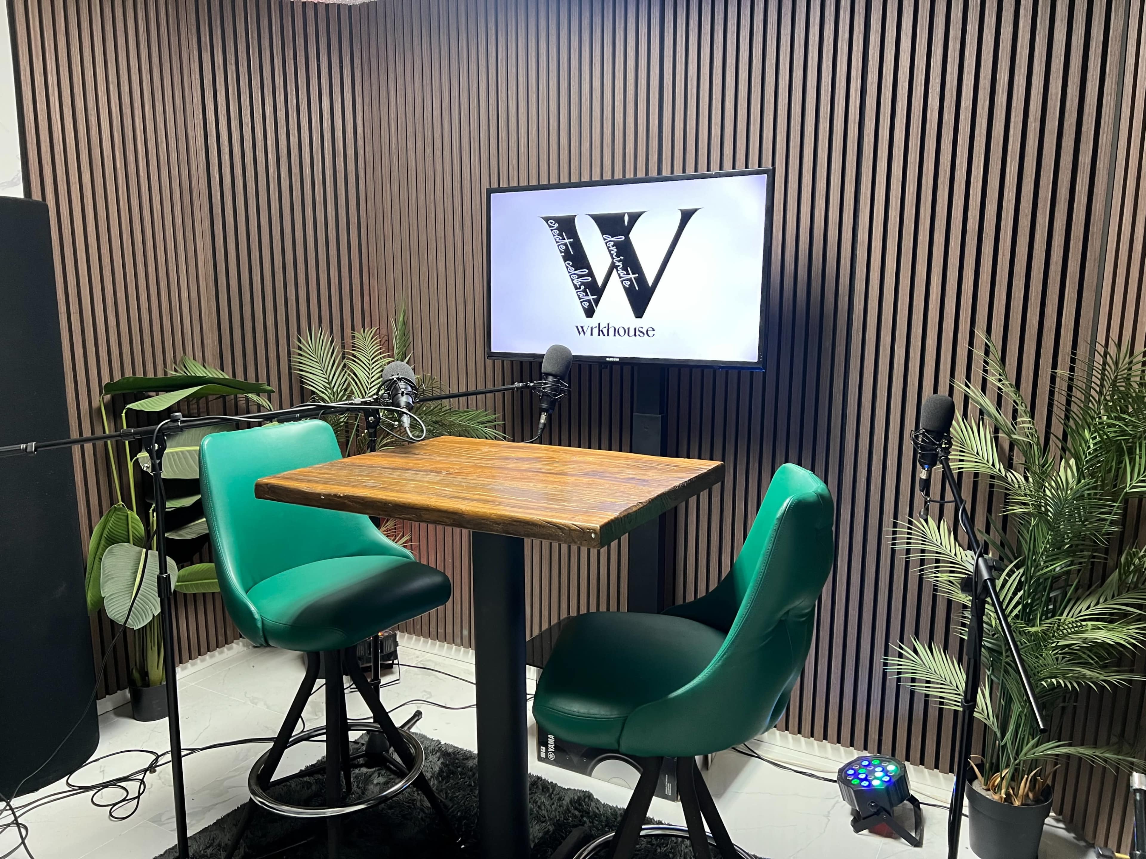 The image shows a podcast recording setup featuring a wooden table, two green chairs, and a television screen displaying the logo "W" against a backdrop of vertical wooden slats and decorative plants.