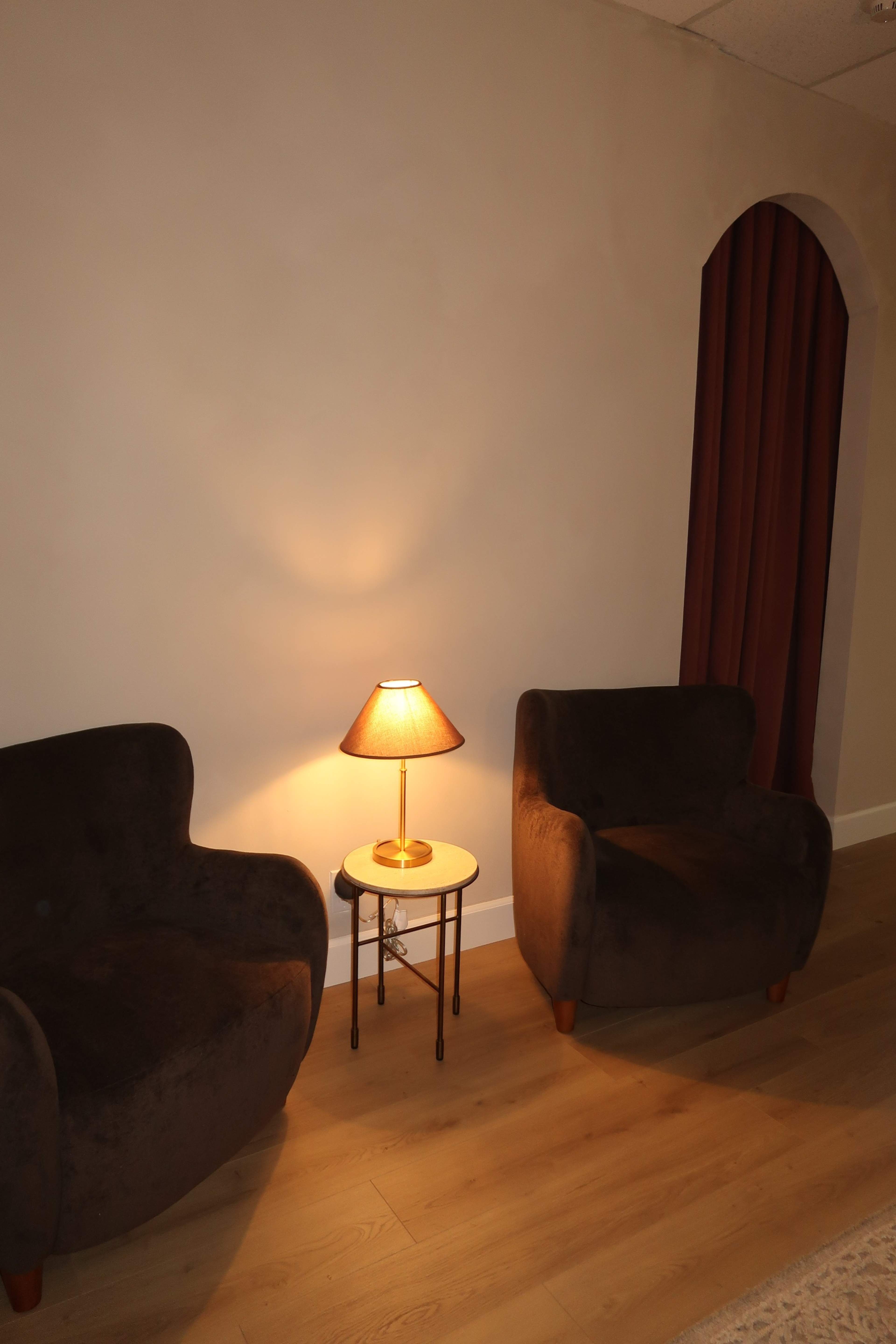 The image shows two dark upholstered chairs next to a small round table with a lamp, set against a plain wall and a curtain.