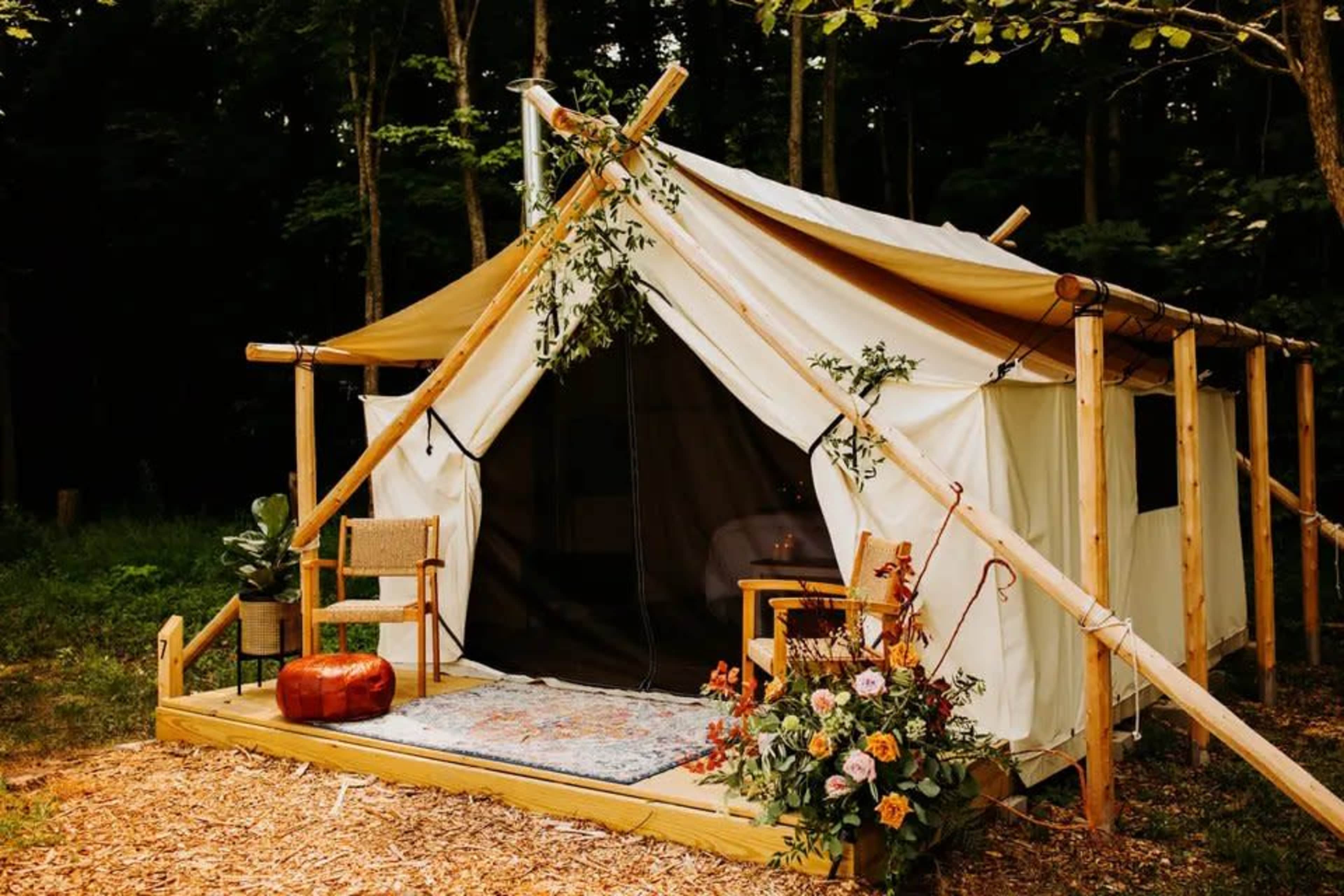 A glamping tent is set in a wooded area, featuring a wooden deck with chairs and decorative plants at the entrance.