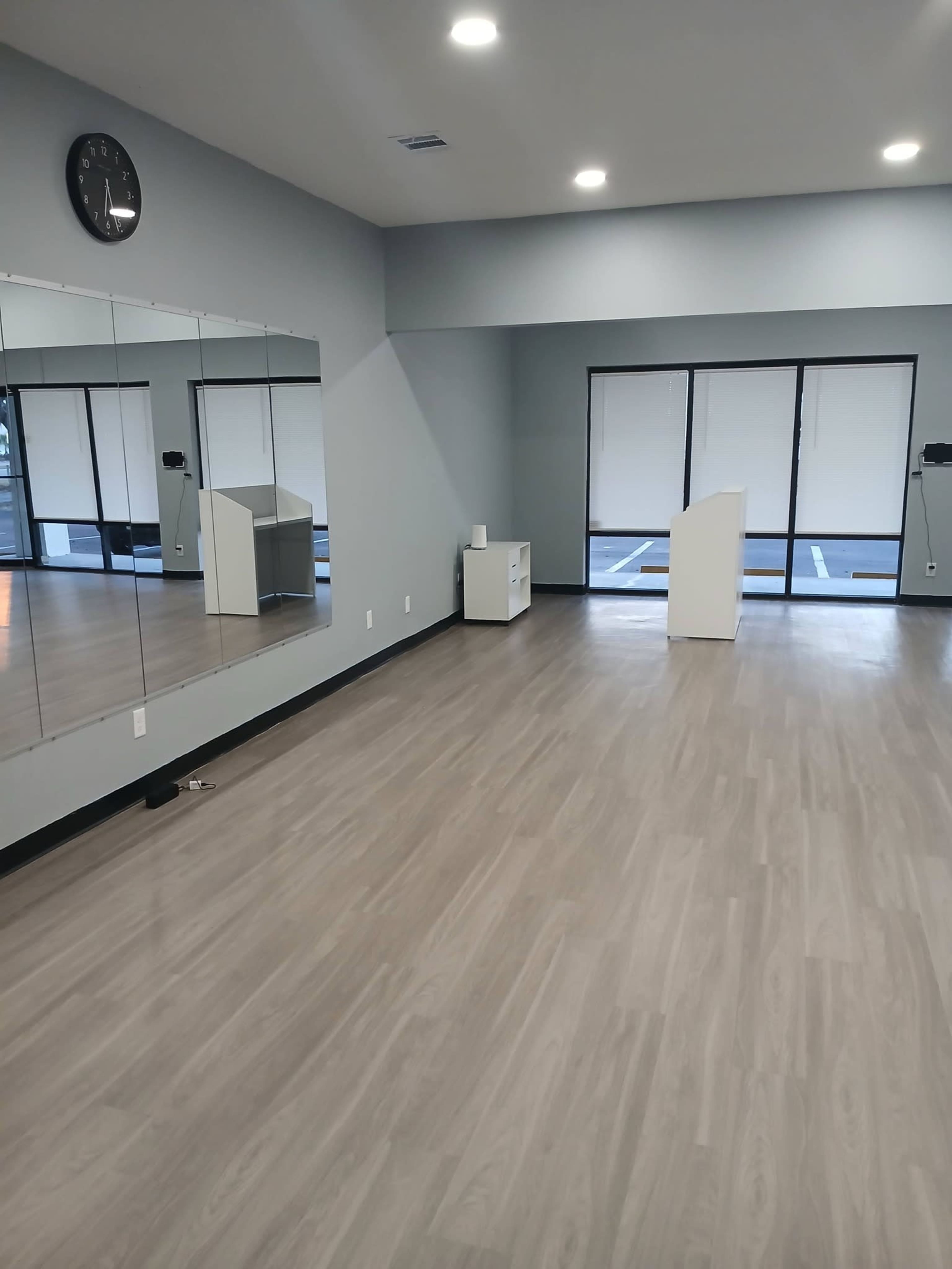 The image shows a spacious, empty interior room with light wood flooring, a large mirror on one wall, and large windows facing outside.