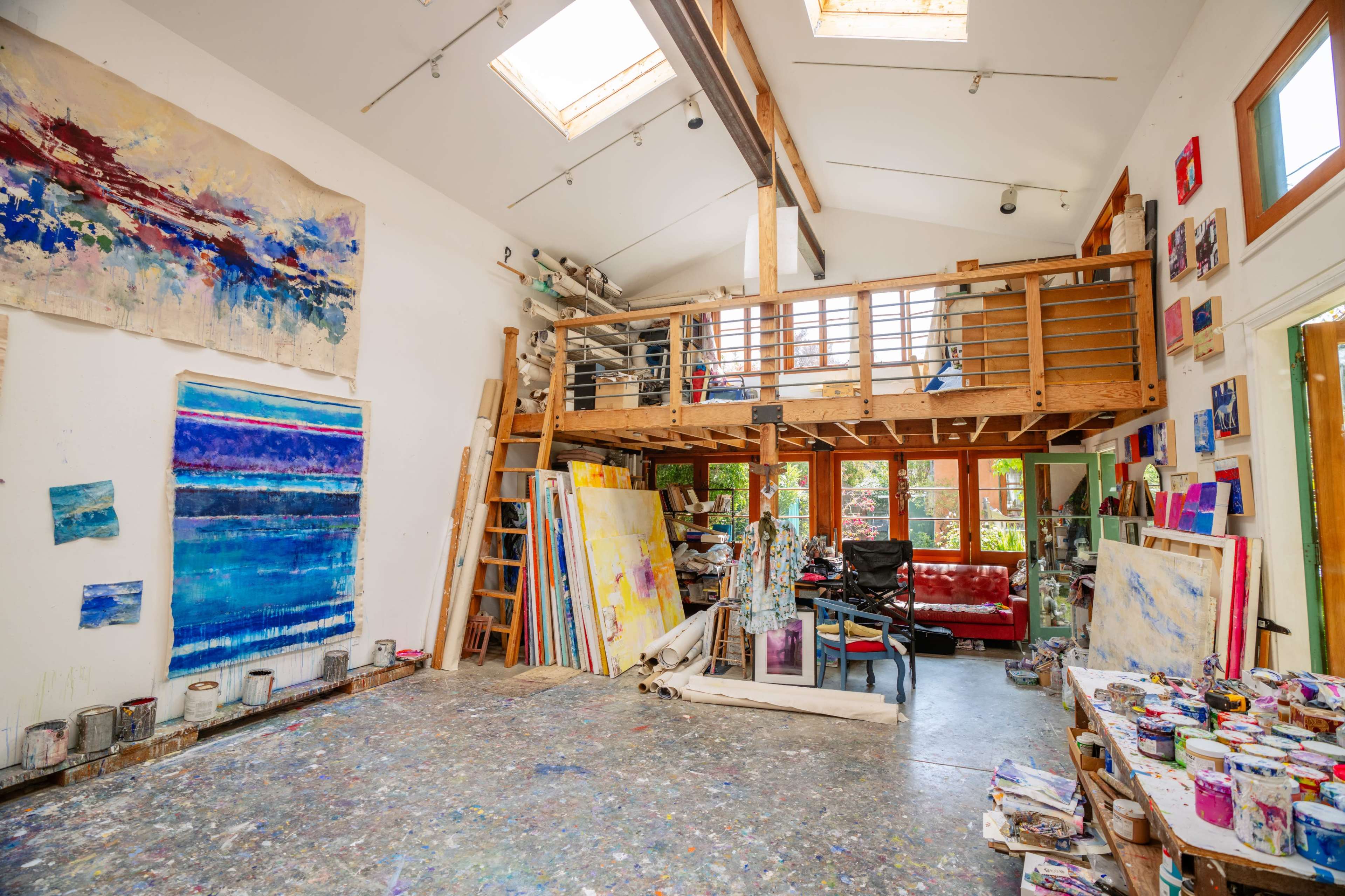A spacious art studio with a wooden loft, featuring vibrant paintings on the walls and a cluttered work area filled with art supplies.