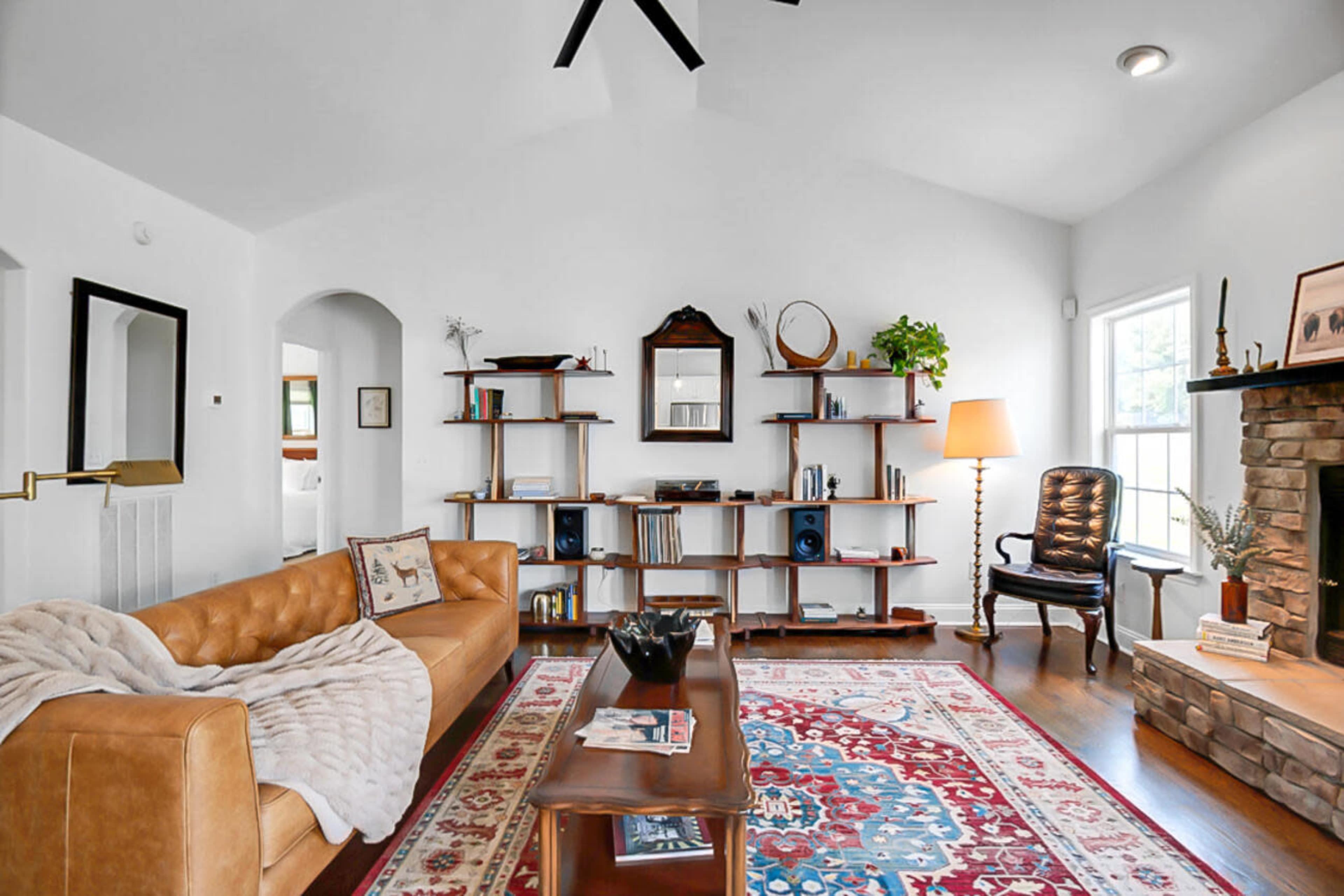 The scene features a cozy living room with a leather sofa, wooden shelves displaying books and decor, a vintage armchair, and a stone fireplace.