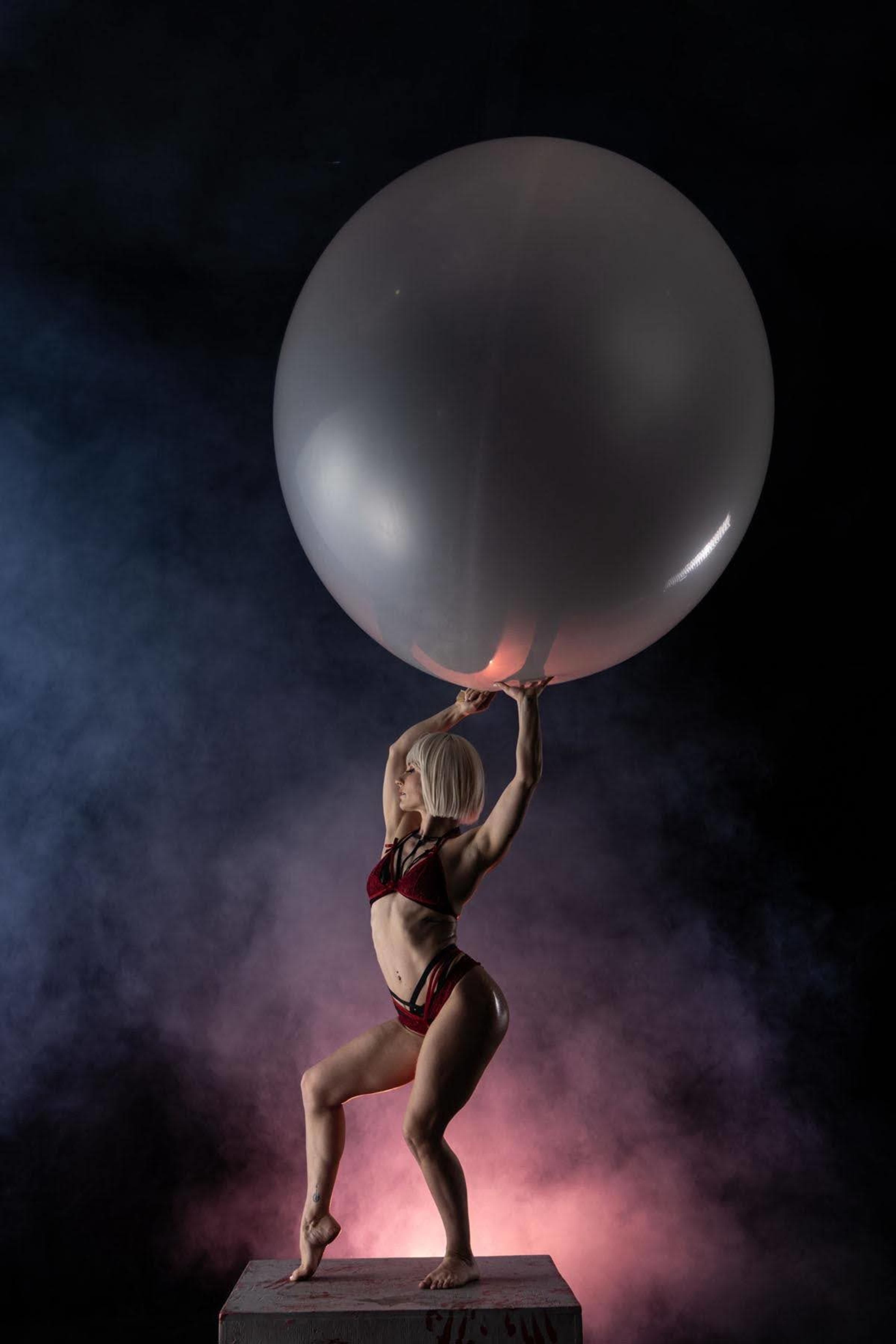 A person in a bikini poses while lifting a large transparent balloon against a smoky, colorful background.