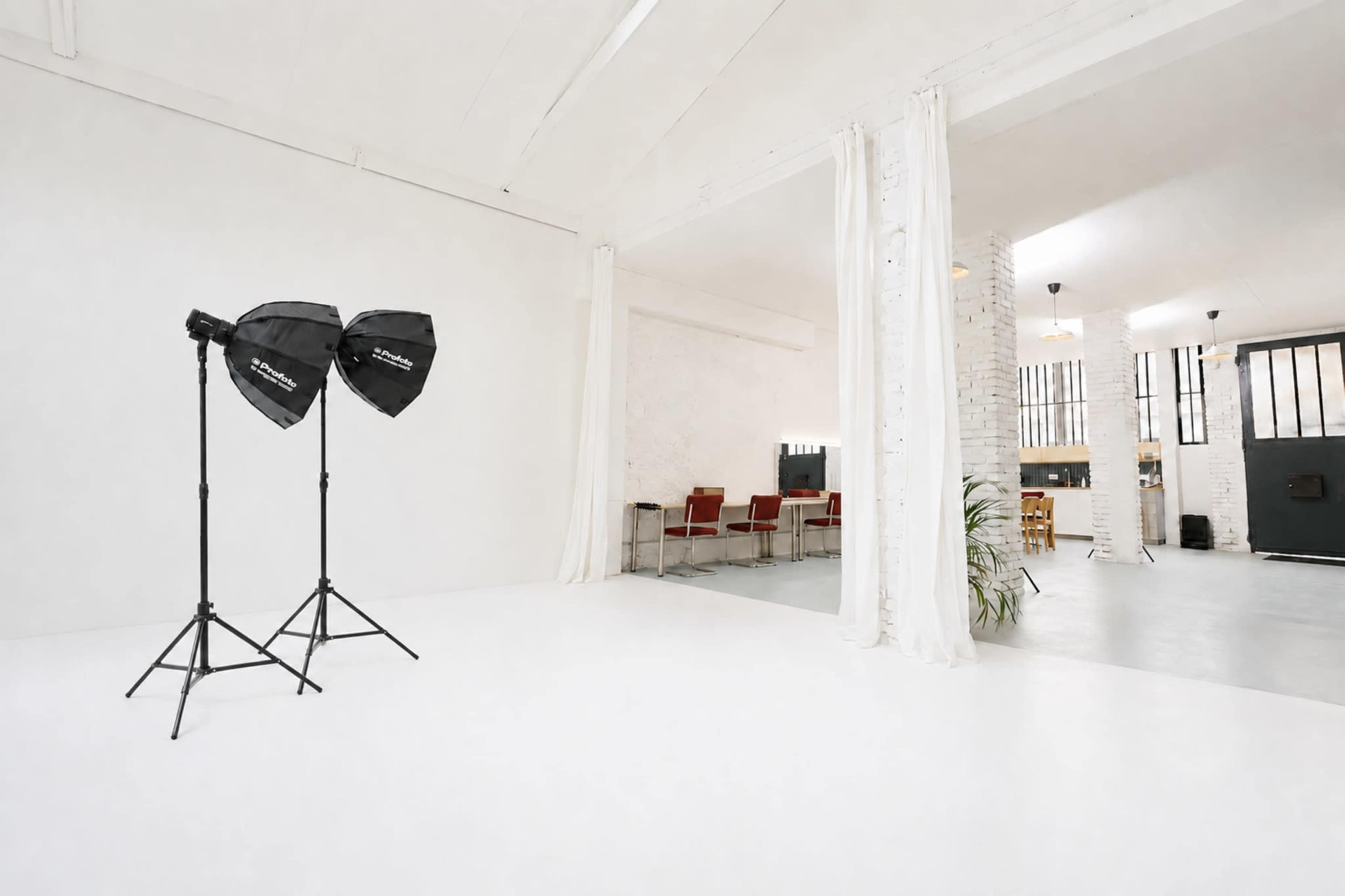 A bright studio space features two light stands with softboxes, white walls, and an area with red chairs and a dining table in the background.