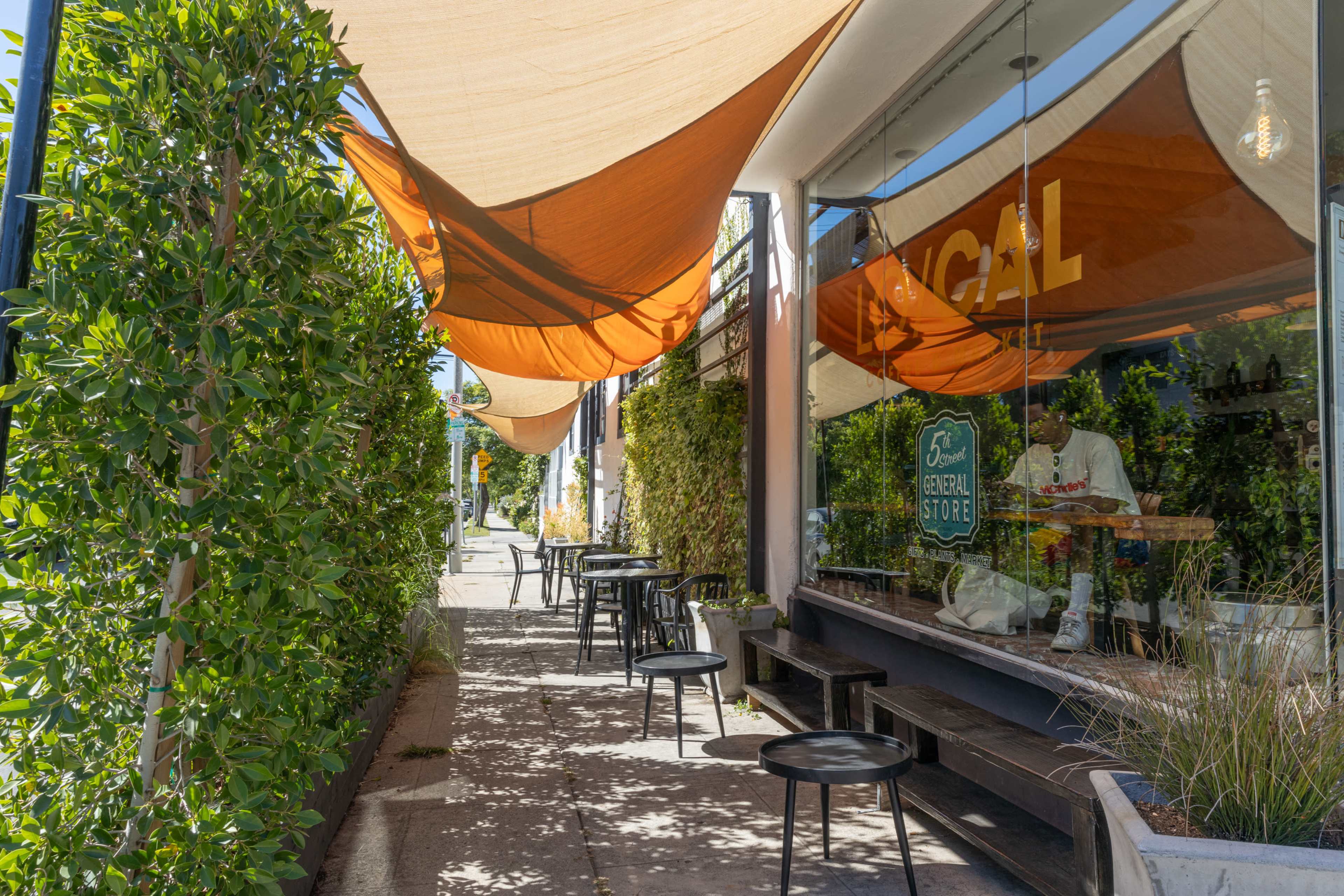 A narrow sidewalk outside a café is lined with black tables and chairs, shaded by orange canopies, with greenery on either side.