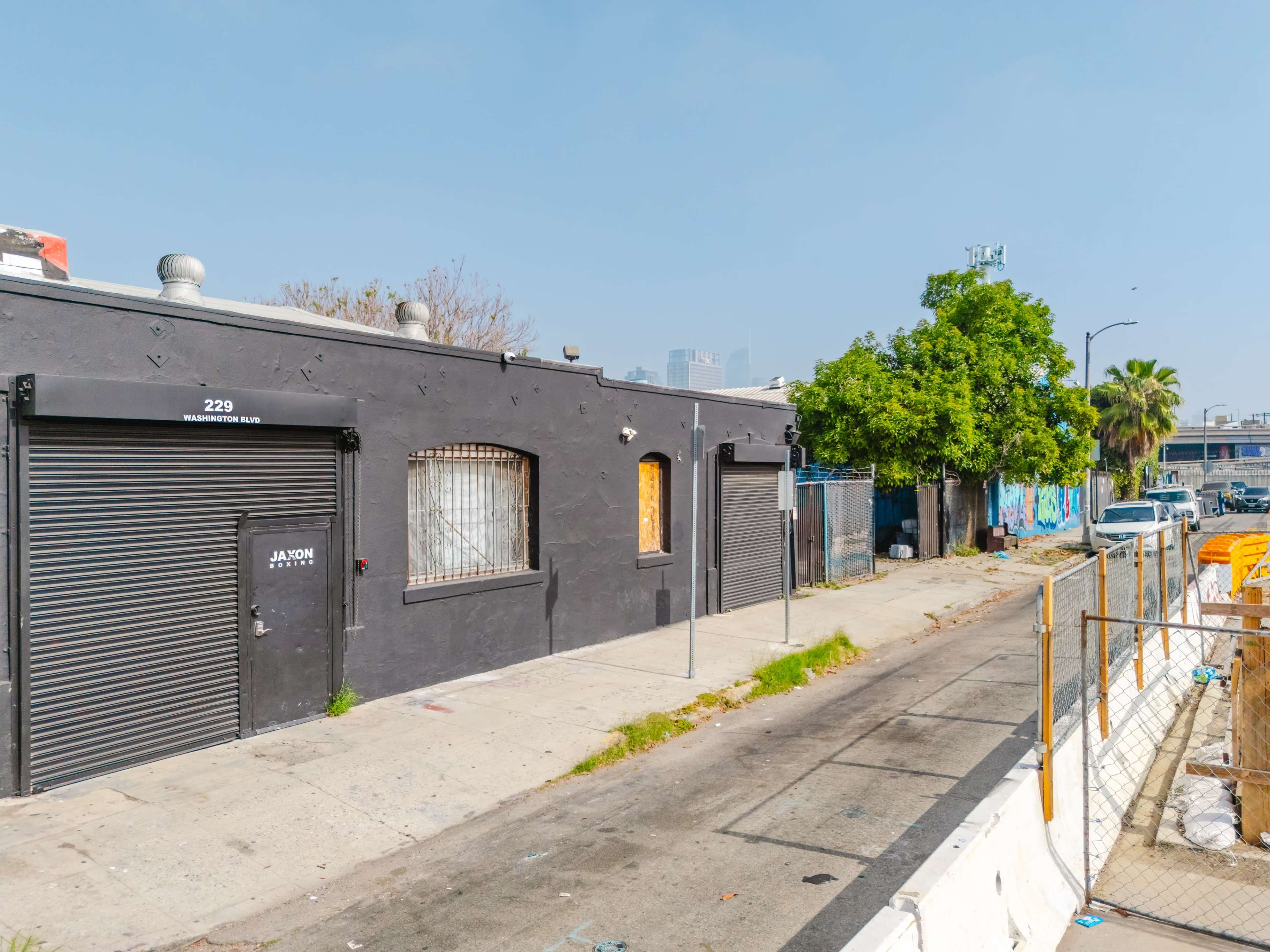The image shows a narrow street lined with a black building featuring closed roll-down doors, graffiti-covered walls, and visible palm trees in the background.