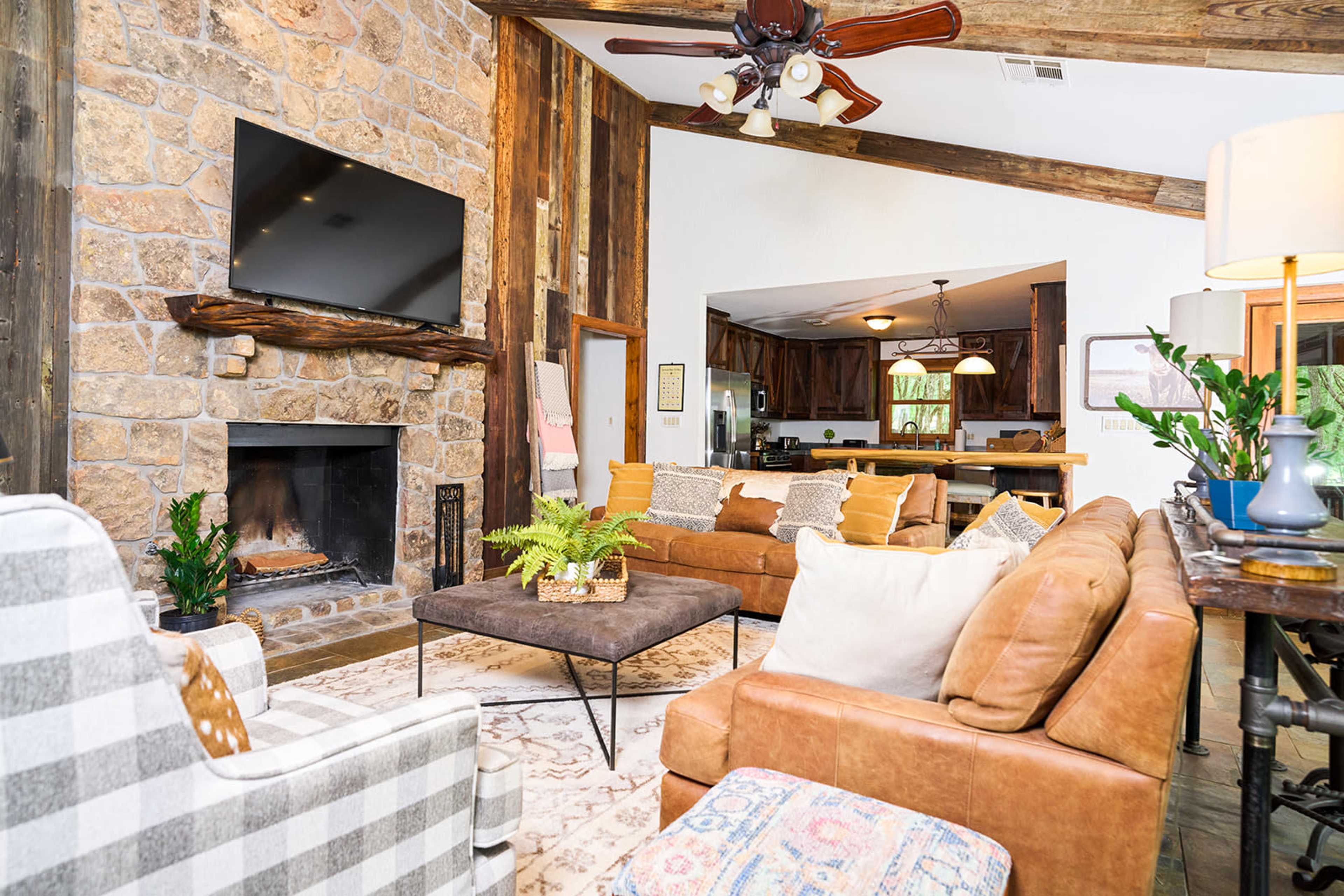 The living room features a stone fireplace, a large flat-screen TV mounted above it, and a mix of seating options including a leather sofa and patterned chairs.
