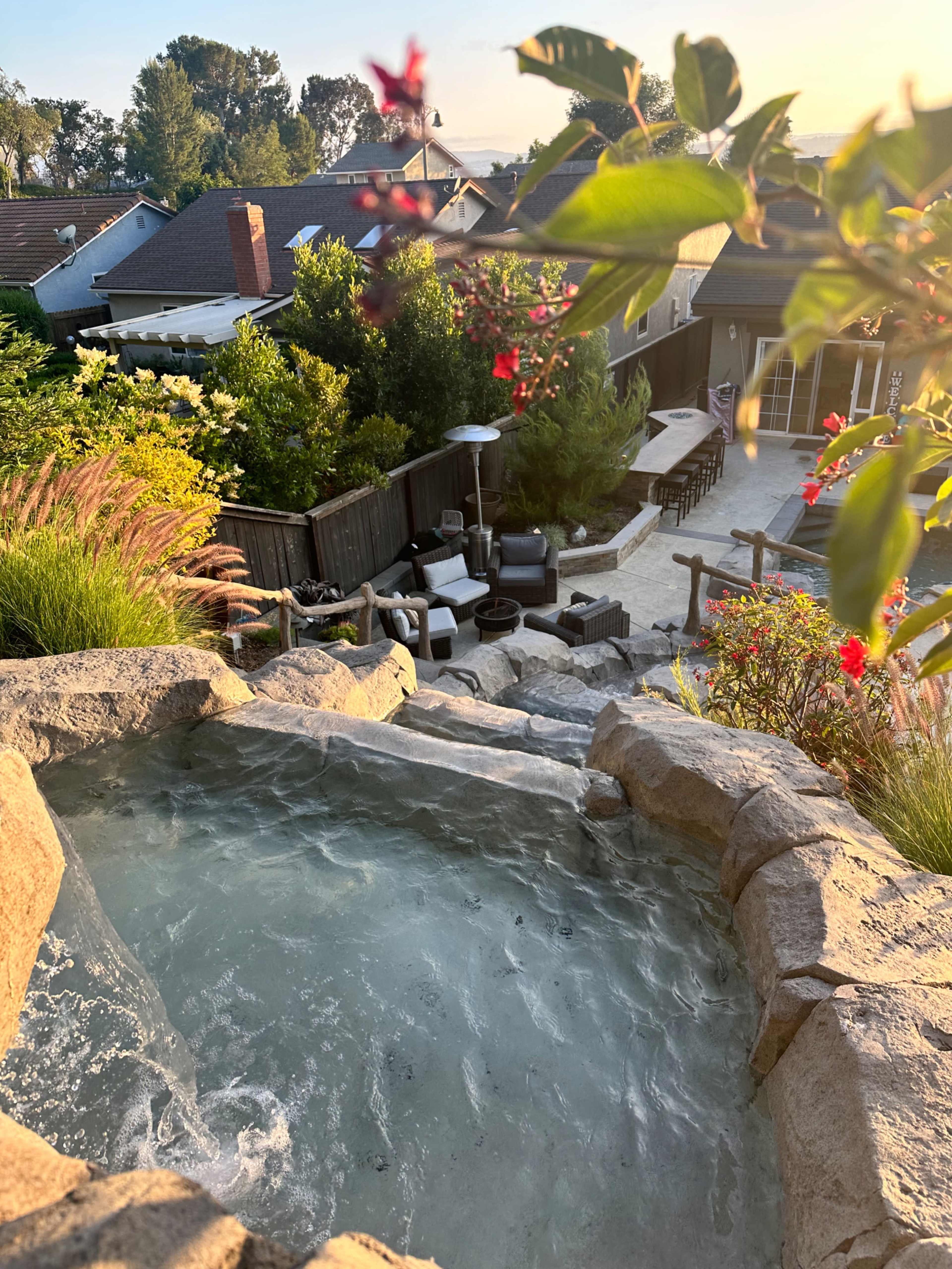 A backyard with a stone hot tub, a patio area with outdoor furniture, and vibrant greenery under a sunset sky.