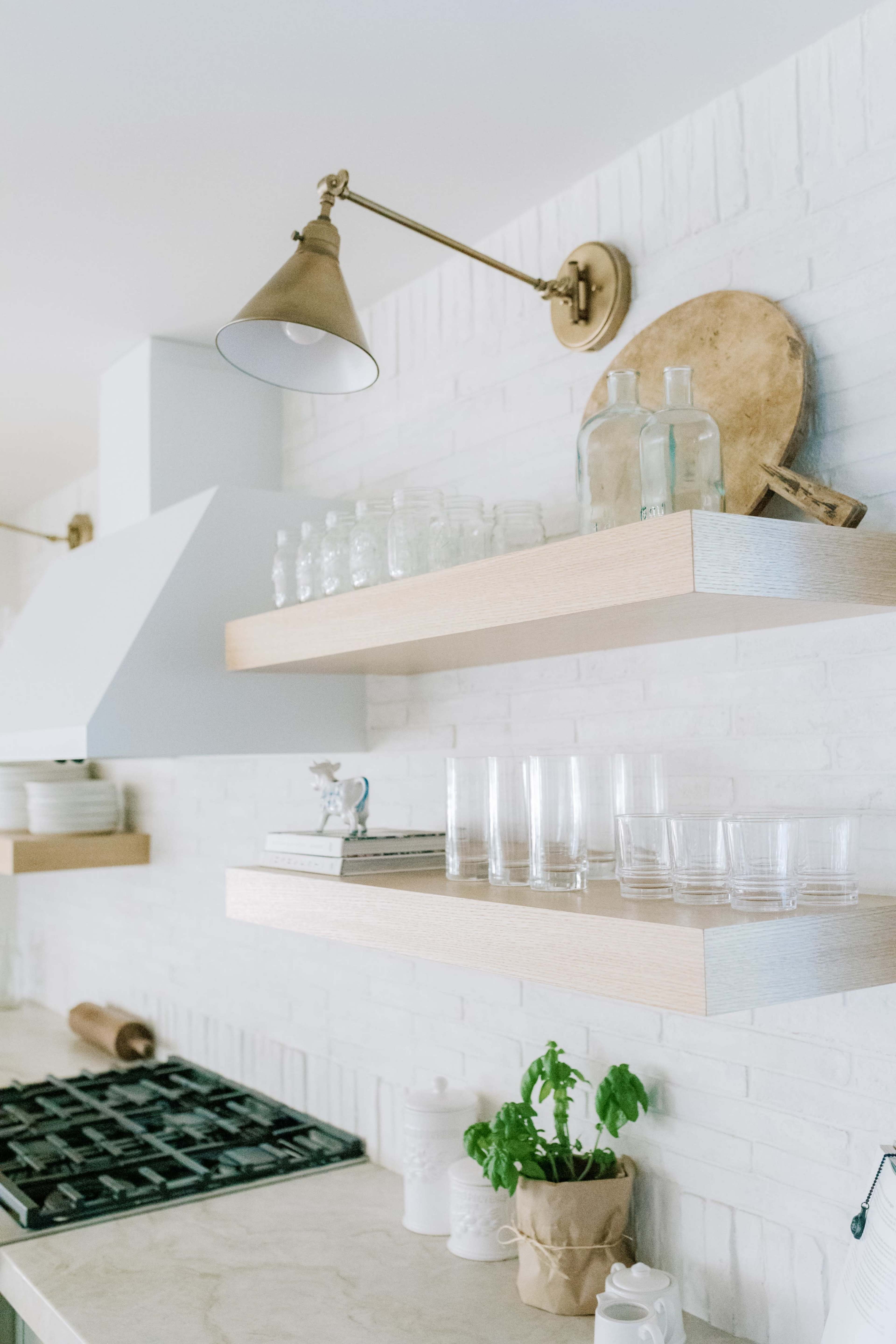 The image shows a modern kitchen with white brick walls, wooden shelves holding glassware and a few decorative items, and a brass wall light above.