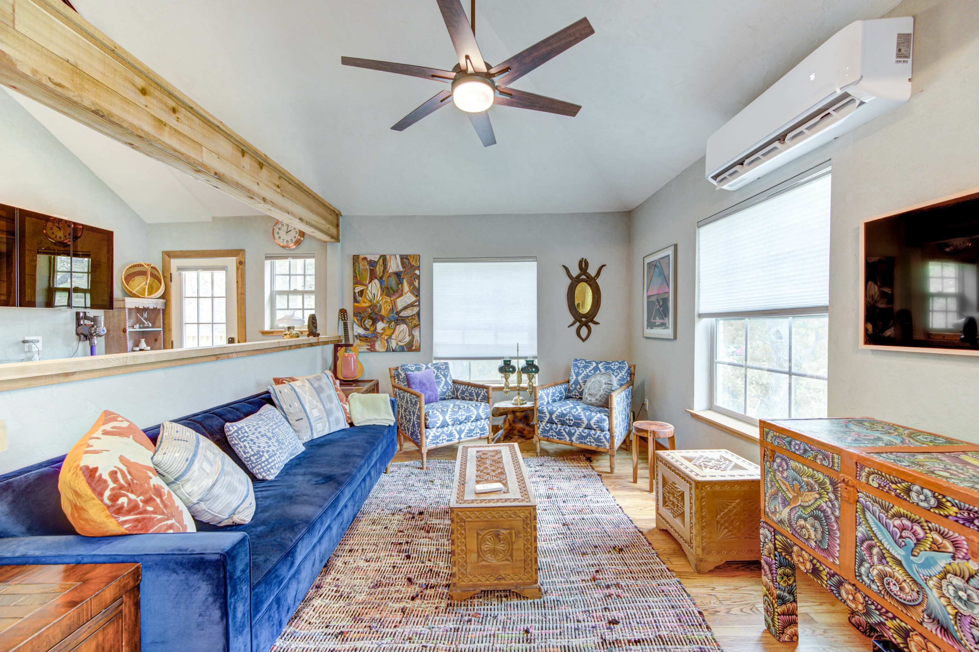 A cozy living room featuring a blue sofa, patterned armchairs, and decorative wooden furniture under a ceiling fan, with natural light streaming through the windows.