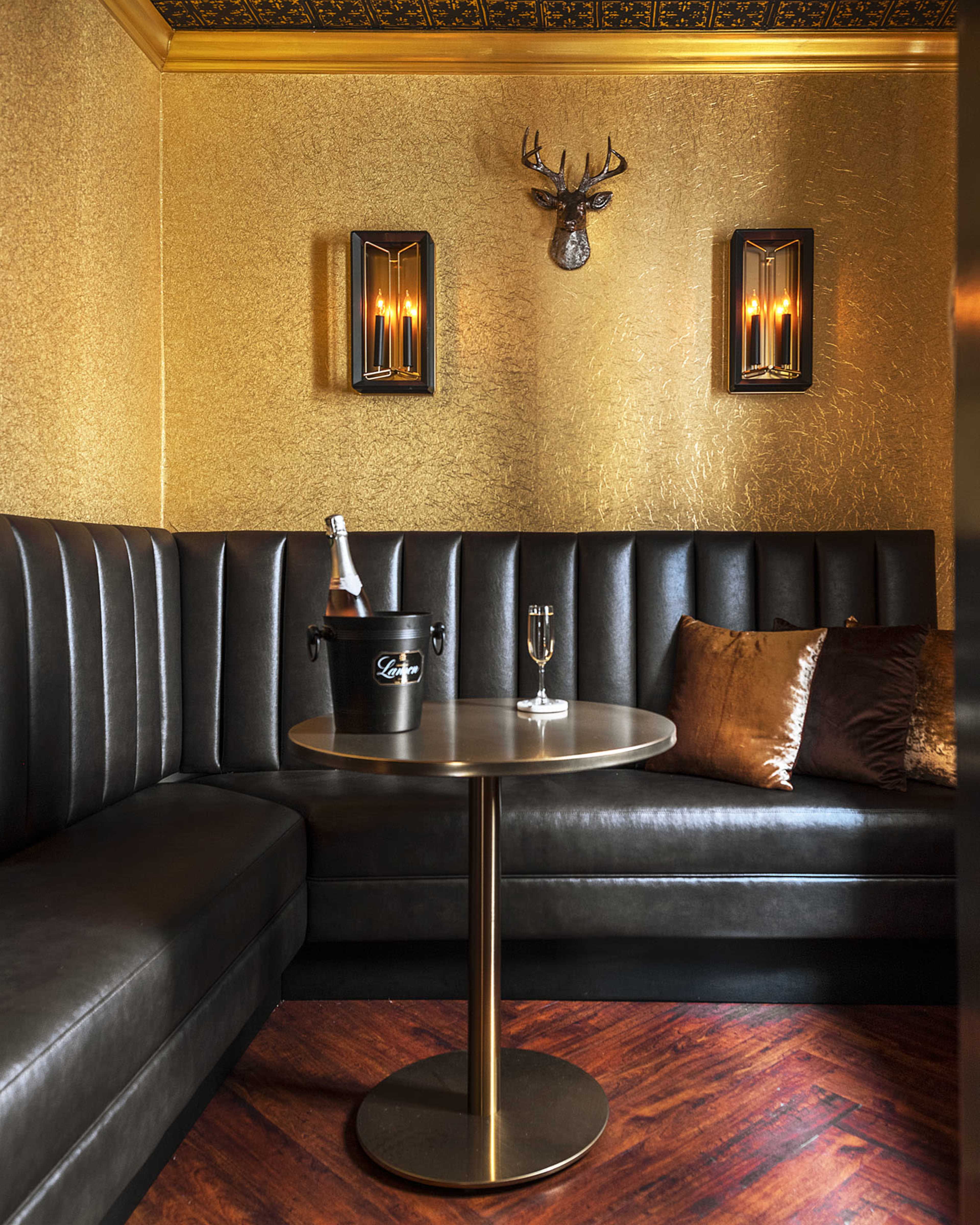 A luxurious seating area with a black leather booth, a round gold table, a bottle of champagne in an ice bucket, and decorative wall sconces in a golden-hued room.