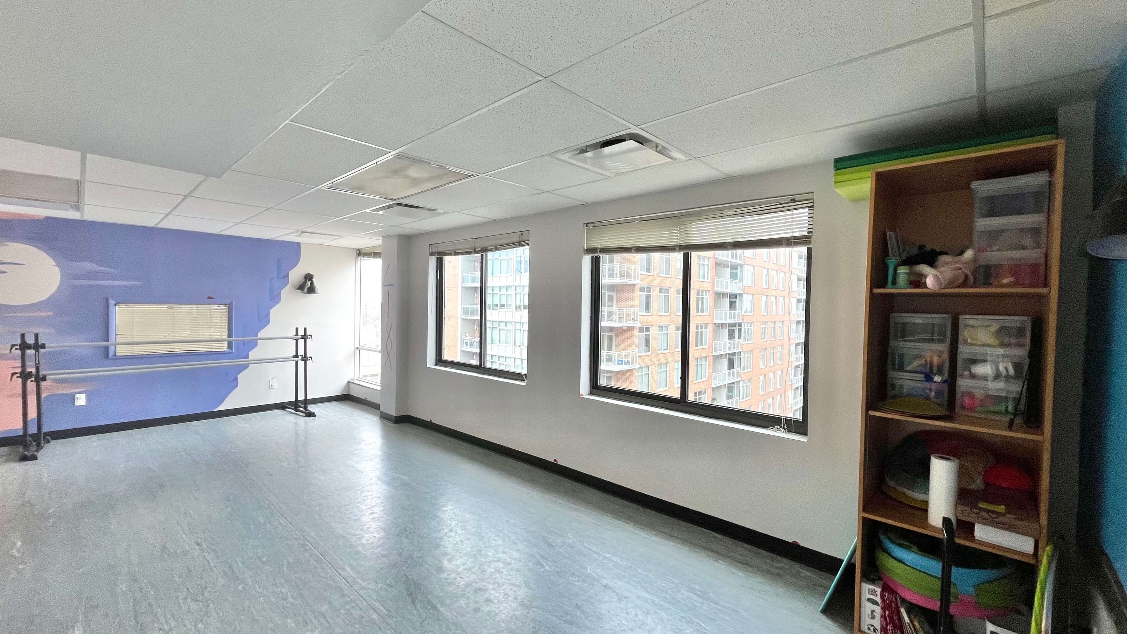 An empty dance studio with large windows, a mirrored wall, and a shelving unit containing various items.