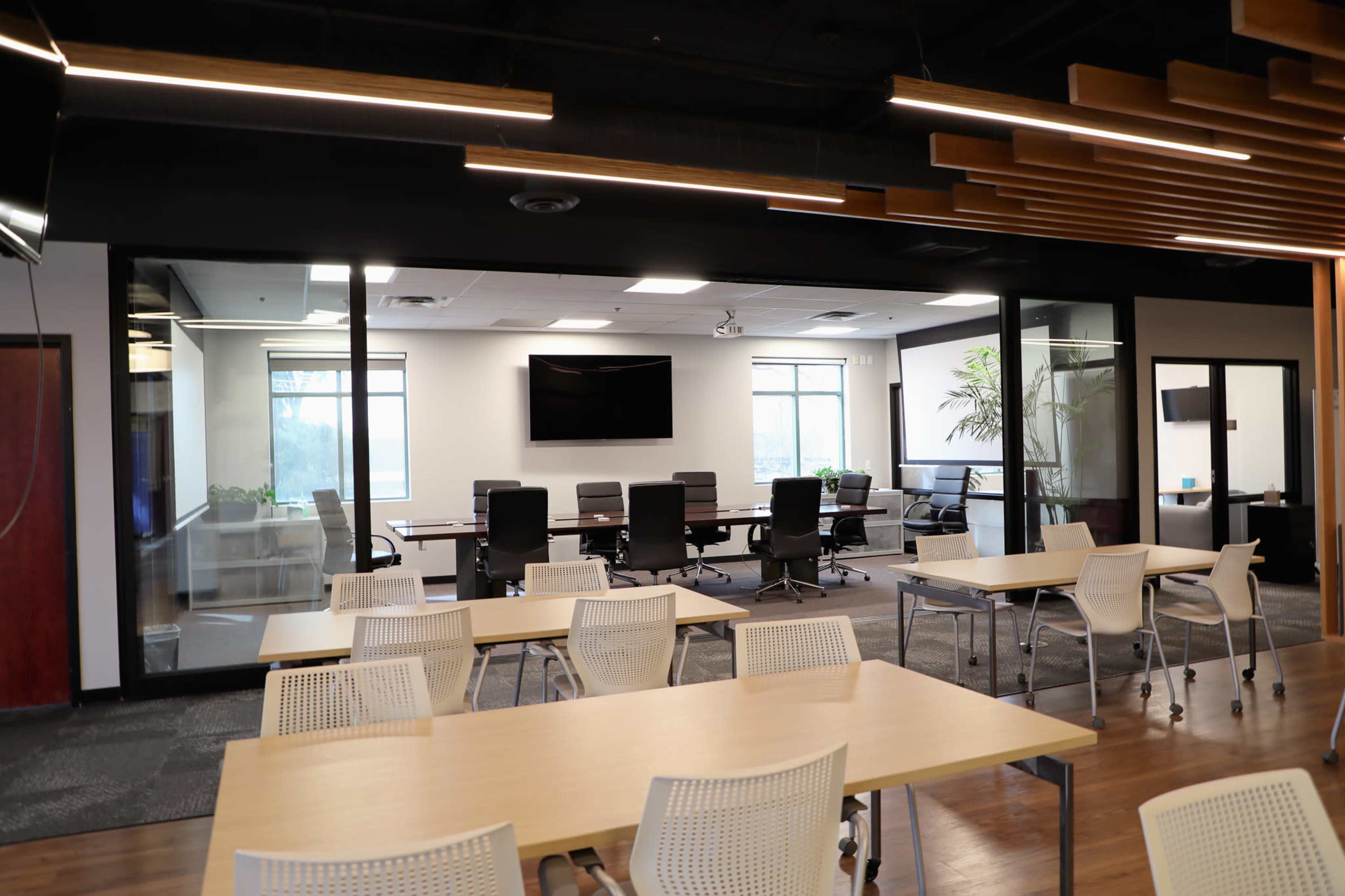 A modern office space features several tables and chairs in the foreground, with a glass-walled conference room visible in the background.